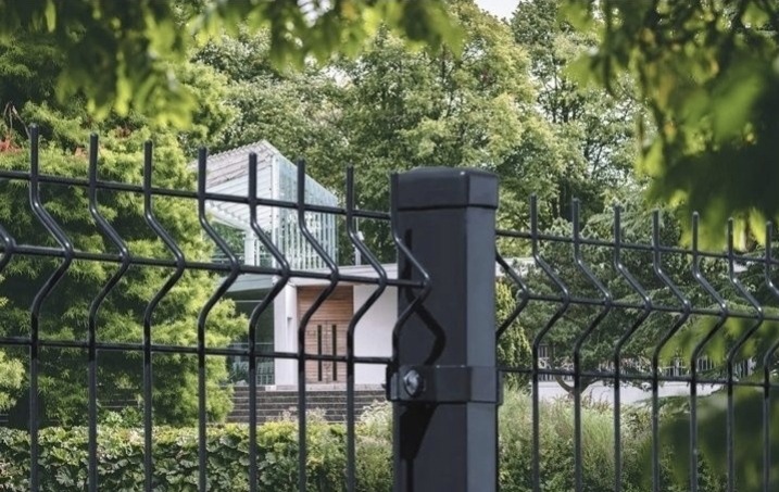 Czarne, panelowe ogrodzenie na tle zieleni i nowoczesnego domu z białymi elementami, widok z bliska, spokojna kolorystyka.