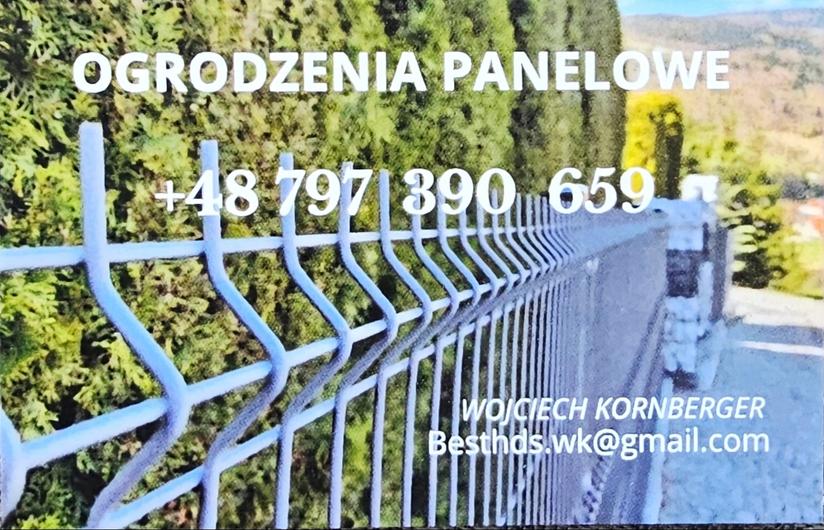 Niebieskie ogrodzenie panelowe z danymi kontaktowymi na tle zieleni i gór. Widok z boku, uwydatniający strukturę paneli i słupków.