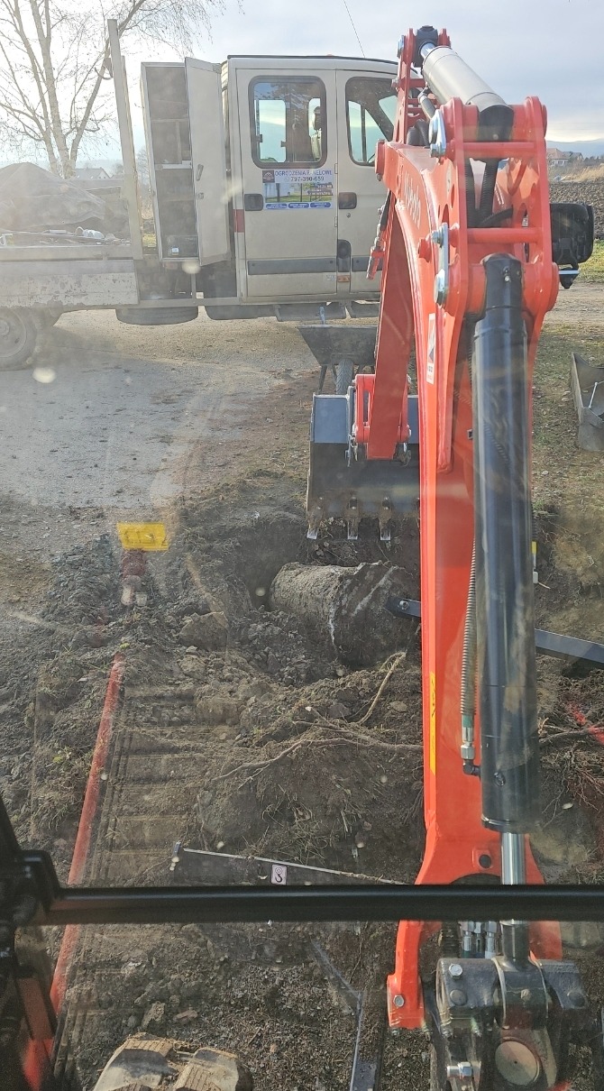 Pomarańczowa koparka w trakcie wykopu w ziemi, widok z kabiny operatora, w tle samochód dostawczy z otwartymi drzwiami i reklamą firmy.