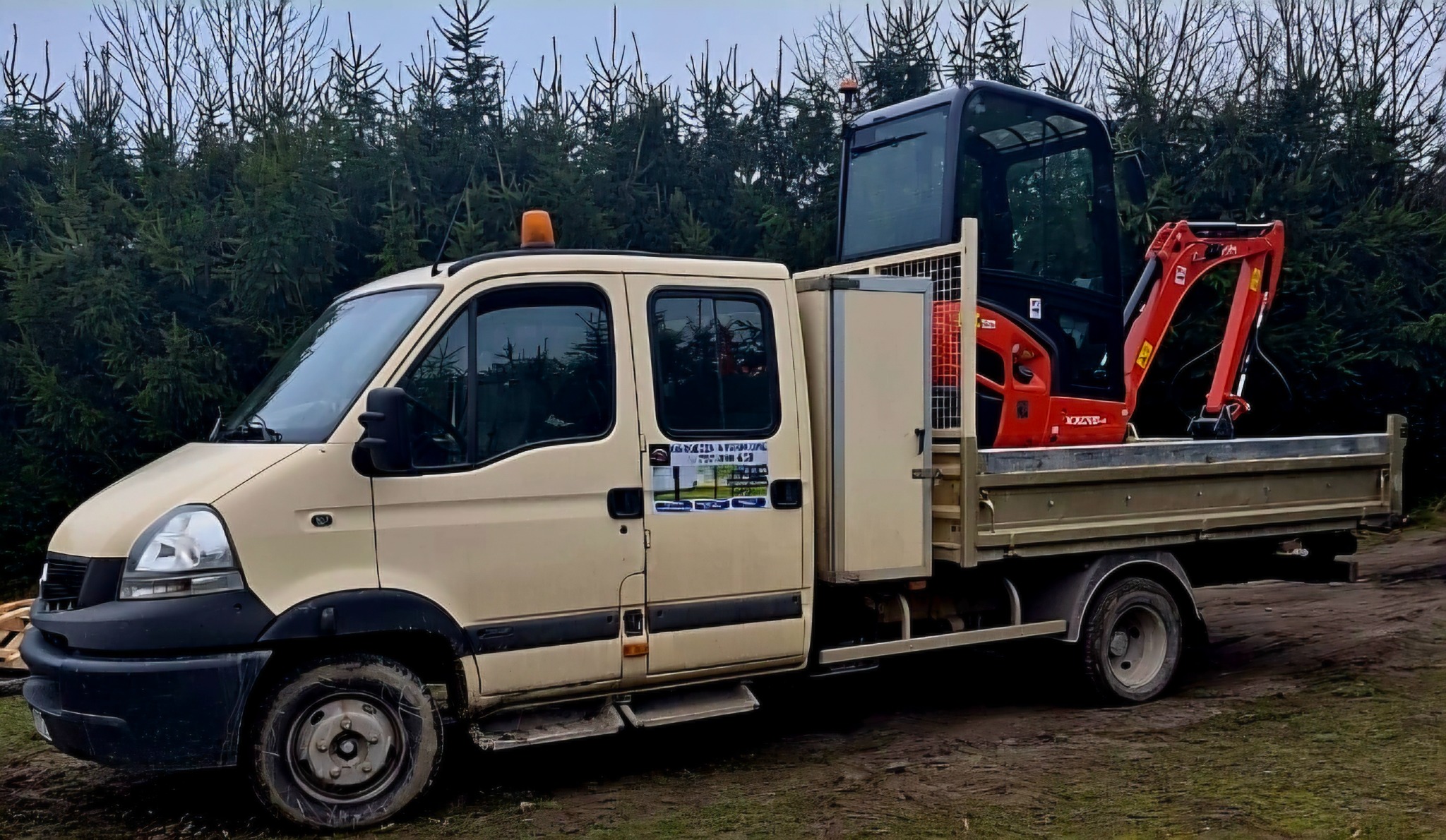 Beżowy samochód dostawczy z czerwoną mini koparką na pace, na tle zielonych drzew iglastych. Widoczna naklejka z danymi firmy na drzwiach.
