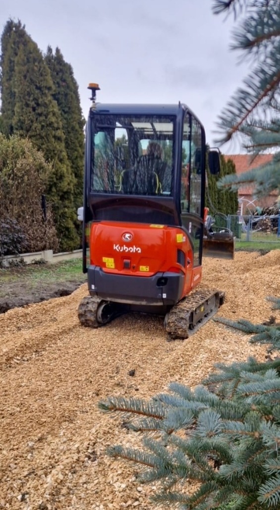 Pomarańczowa minikoparka Kubota na terenie pokrytym drobnym żwirem, widoczne świeżo wykopane rowy i zieleń w tle. Prace ziemne w ogrodzie.