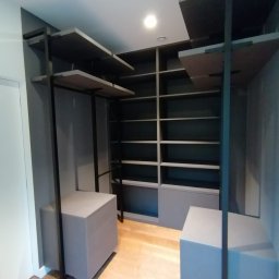 Yurii Pakenas - Nowoczesna garderoba w odcieniach szarości z drewnianą podłogą, obejmująca półki, szafki i czarne metalowe ramy.