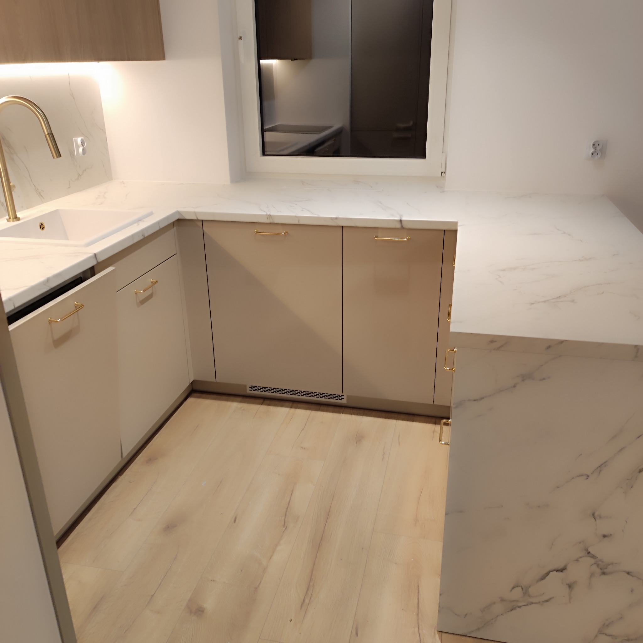 Jasna kuchnia z beżowymi frontami szafek, złotymi uchwytami i blatem imitującym marmur, widok na okno i fragment podłogi z paneli.