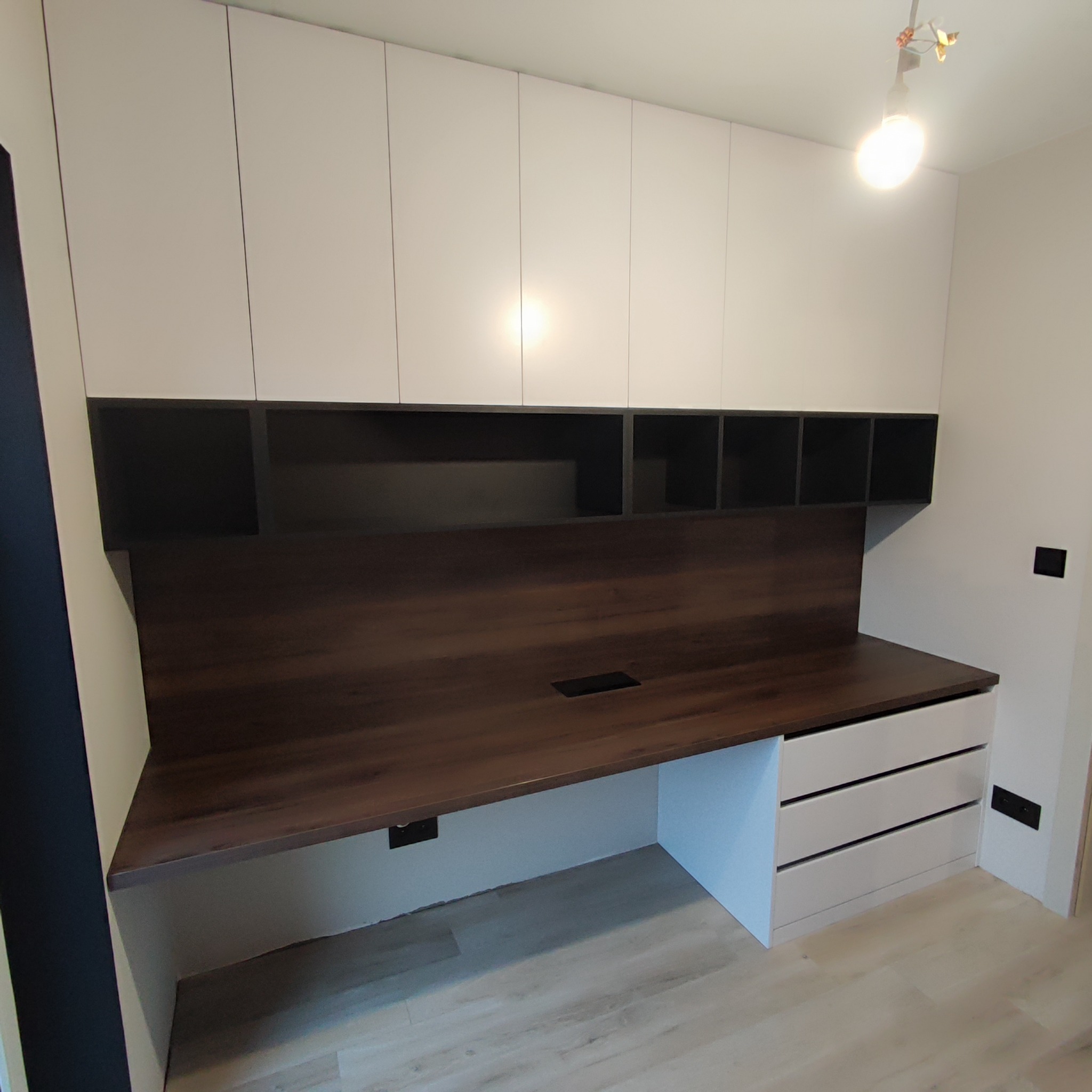 Nowoczesne biurko z blatem drewnopodobnym i białymi szafkami wiszącymi, w tym szafki z otwartymi półkami, z widocznym otworem na kable w blacie.