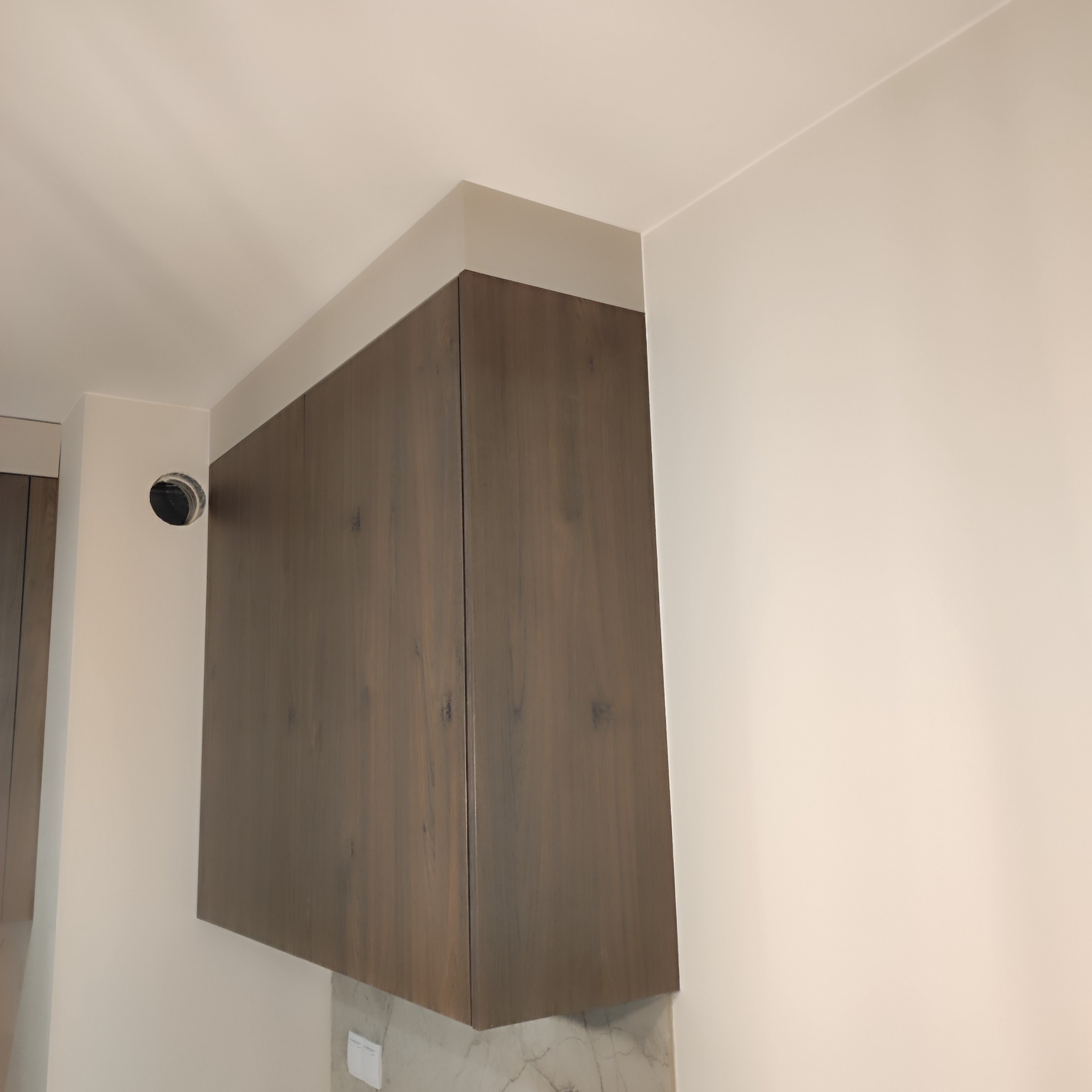 Zamontowana szafka kuchenna wisząca z ciemnego drewna z widocznym fragmentem blatu z marmurowym wzorem oraz otworem w ścianie na przewody.