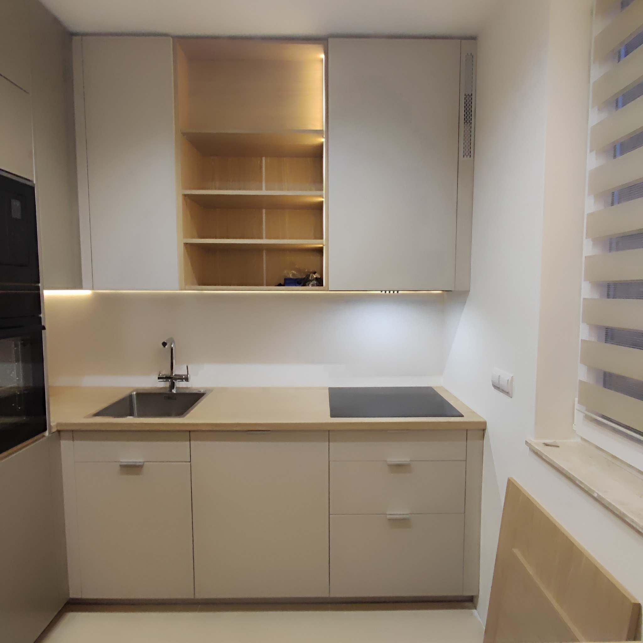 Jasna, nowoczesna kuchnia z górnymi szafkami z otwartymi półkami podświetlonymi od wewnątrz, minimalistyczny zlew i płyta indukcyjna wkomponowane w jasny blat.