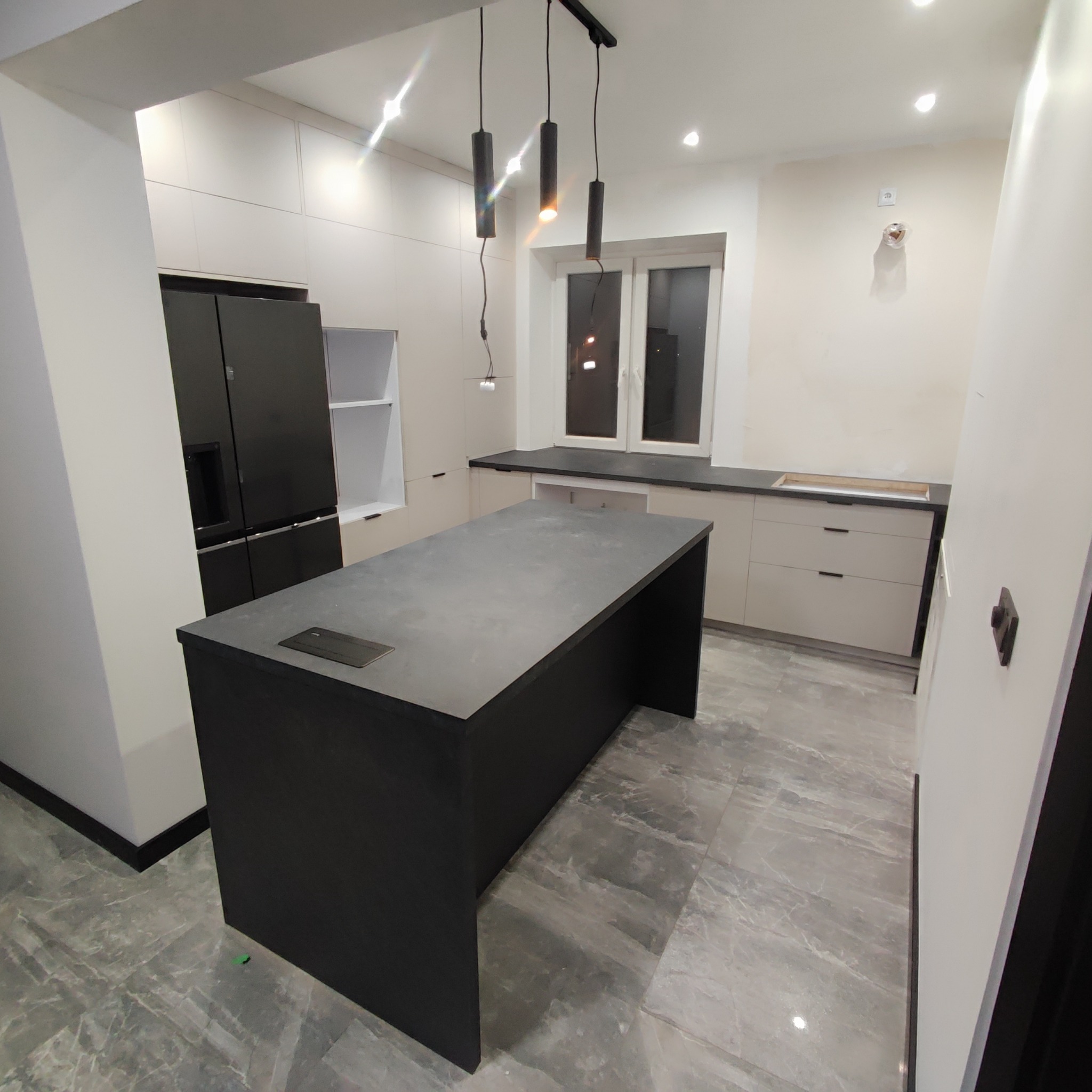 Nowoczesna kuchnia z wyspą, jasnymi szafkami i czarnym blatem, oświetlona trzema wiszącymi lampami.