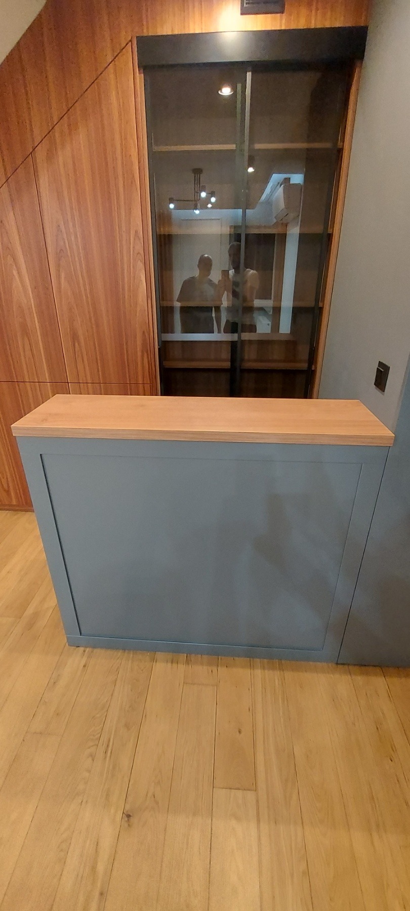 Szara zabudowa kuchenna z drewnianym blatem, widoczna na tle drewnianej szafy i przeszklonej witryny, z odbiciem dwóch osób w szybie.