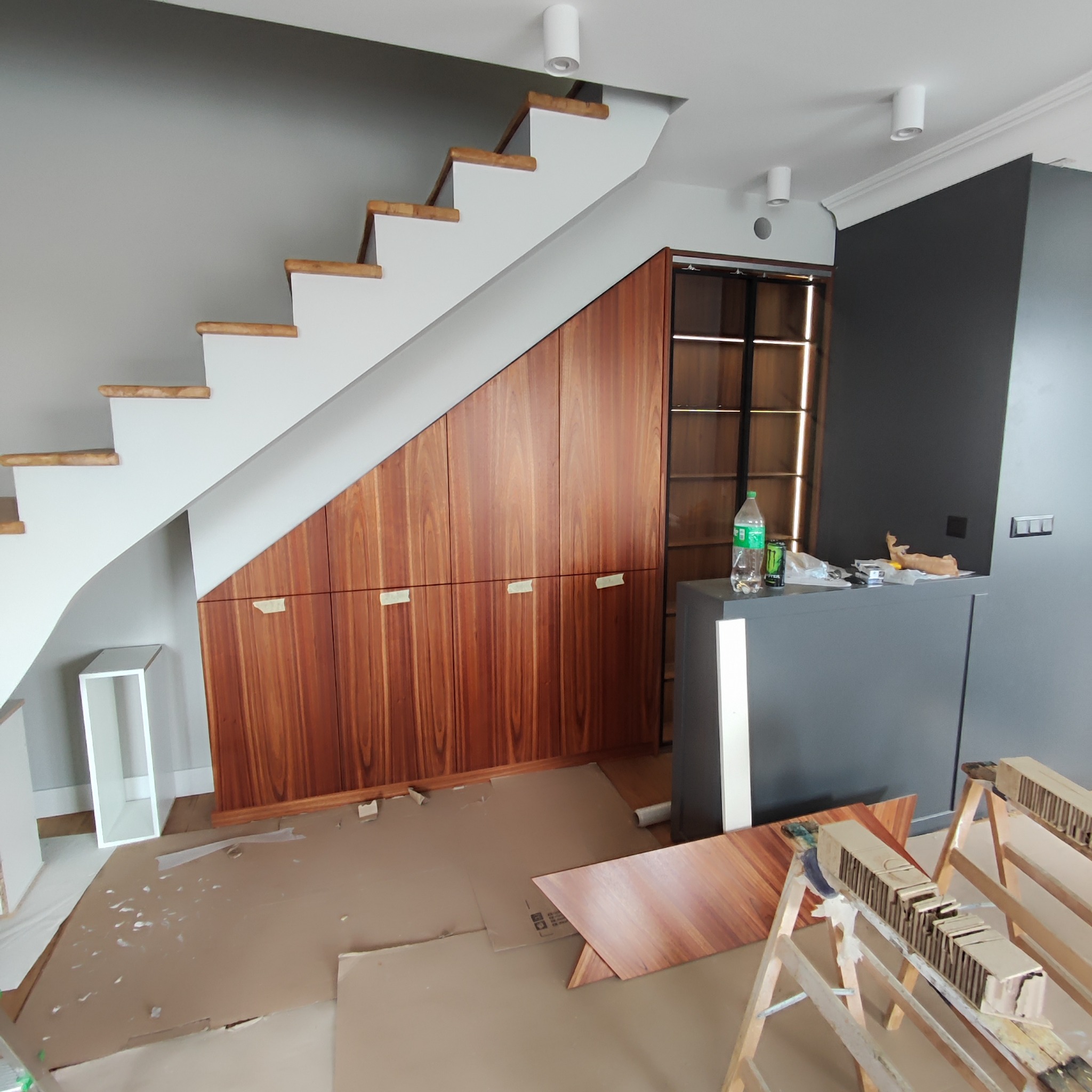 Montaż szafy wnękowej z drewna pod schodami, widoczne elementy na rusztowaniu i kartony zabezpieczające podłogę.
