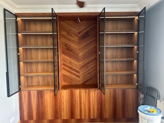 Wysoka zabudowa meblowa z forniru w ciepłych odcieniach brązu, z centralną wnęką o wzorze jodełki i przeszklonymi szafkami po bokach, podświetlonymi od wewnątrz.