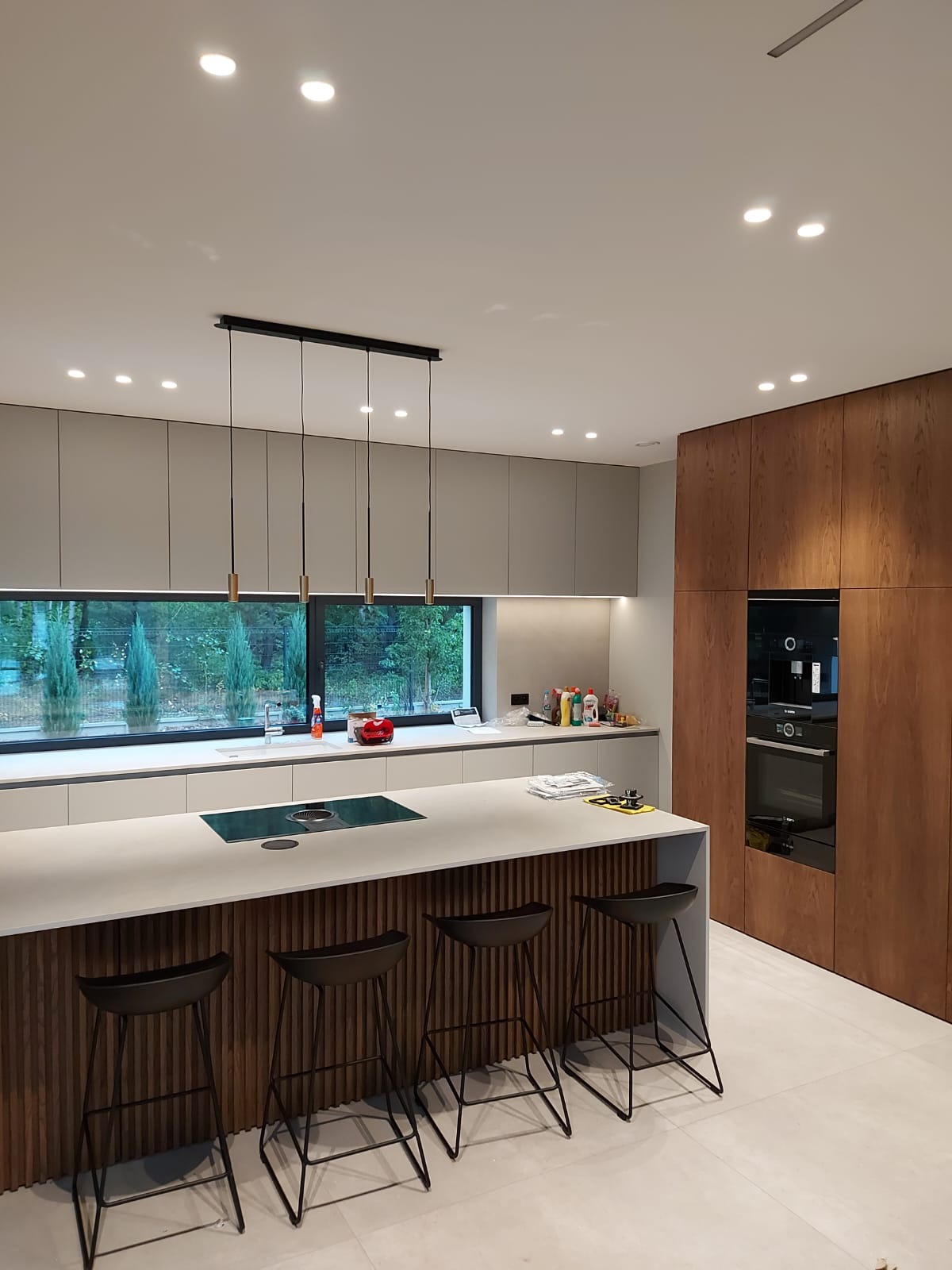 Nowoczesna kuchnia z wyspą, szare szafki wiszące, drewniana zabudowa z piekarnikiem, cztery czarne hokery przy wyspie z ryflowanym frontem, długie okno z widokiem na zieleń.