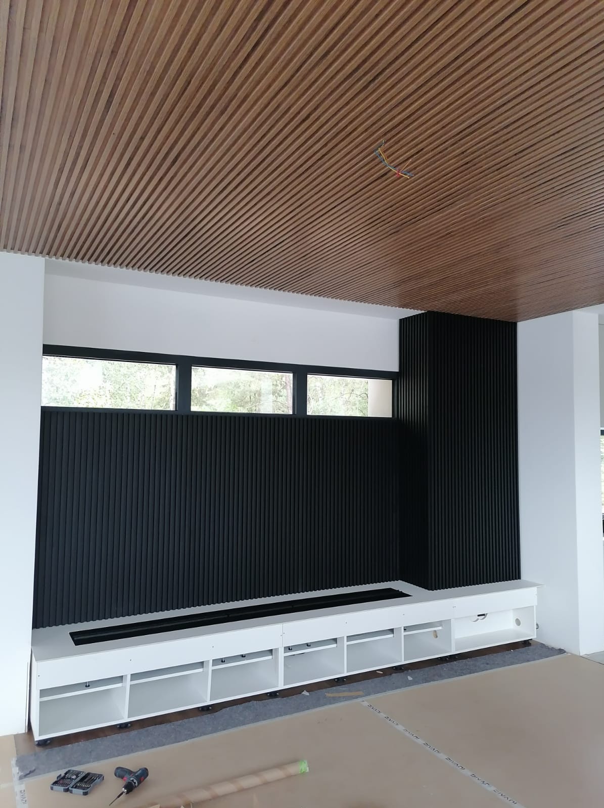 Montaż zabudowy ściany z czarnych paneli lamelowych i białej szafki RTV z otwartymi półkami, widoczne narzędzia i przewody wystające z sufitu.