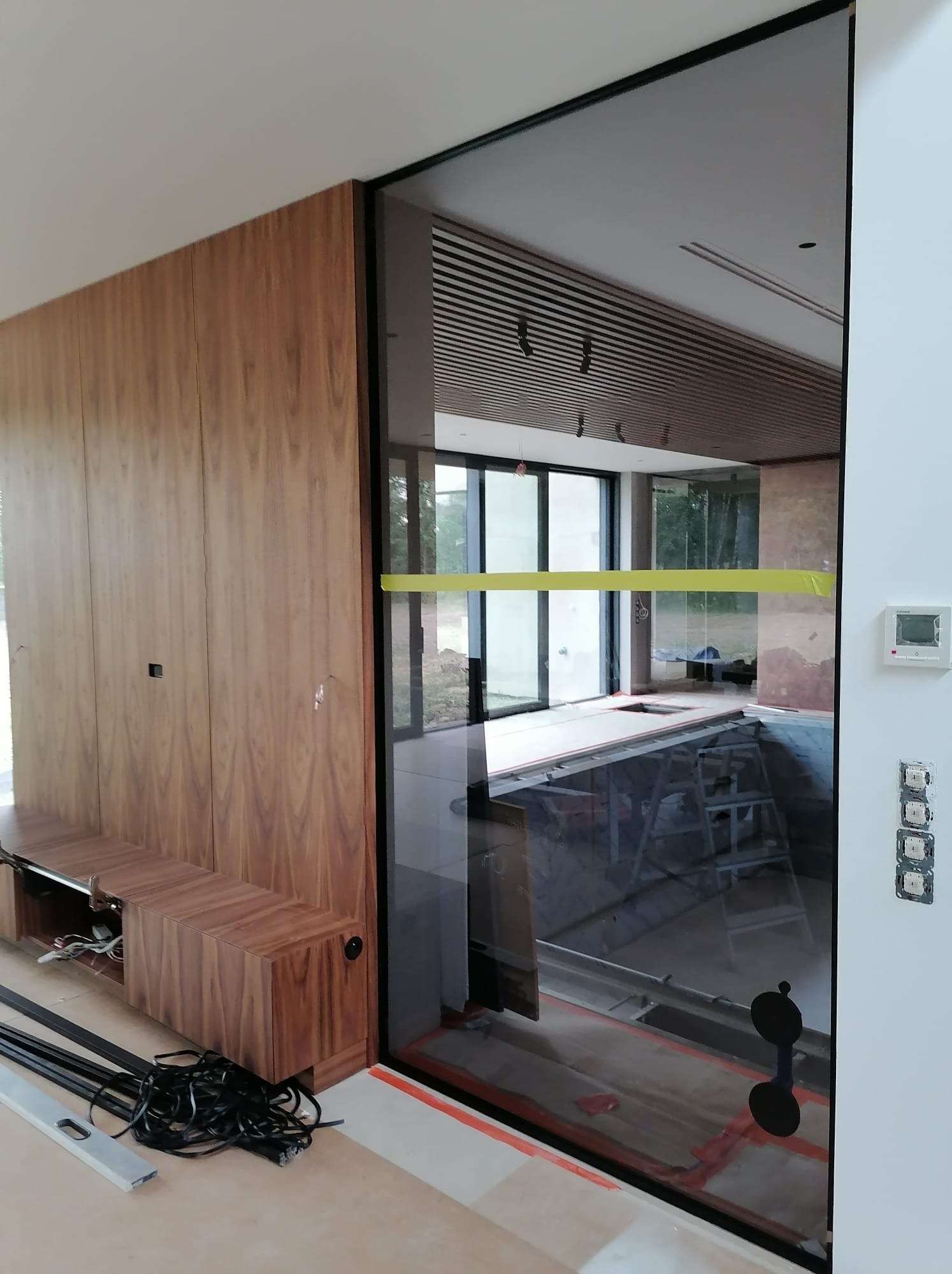Montaż szklanej ściany działowej z czarną ramą, widok na wnętrze w trakcie wykończenia, drewniana zabudowa na ścianie z widocznymi przewodami i poziomica.