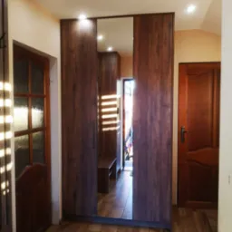 Wnętrze przedpokoju z dużą szafą wnękową z przesuwnymi drzwiami, z lustrem na środkowych drzwiach, odbijającym dalszą część korytarza i światło słoneczne wpadające przez okno.