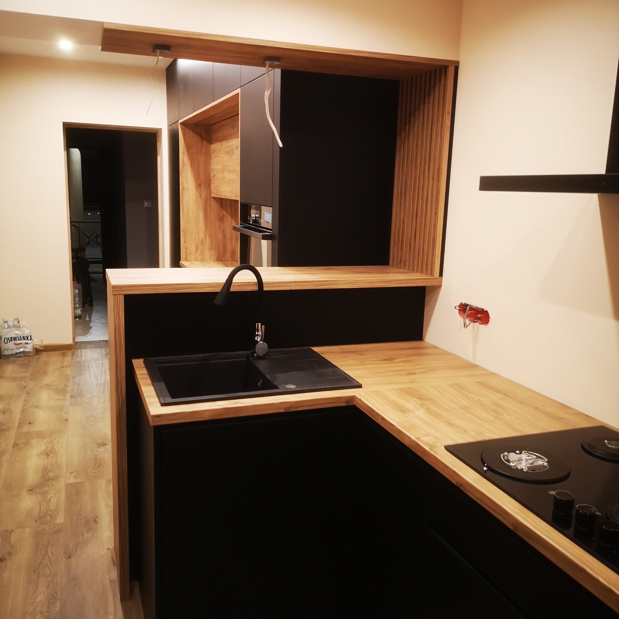 Nowoczesna kuchnia z czarnymi frontami i drewnianymi blatami, widoczny zlew, płyta grzewcza i otwarte półki z drewnianymi panelami, w tle butelki wody mineralnej i niedokończone podłączenie...