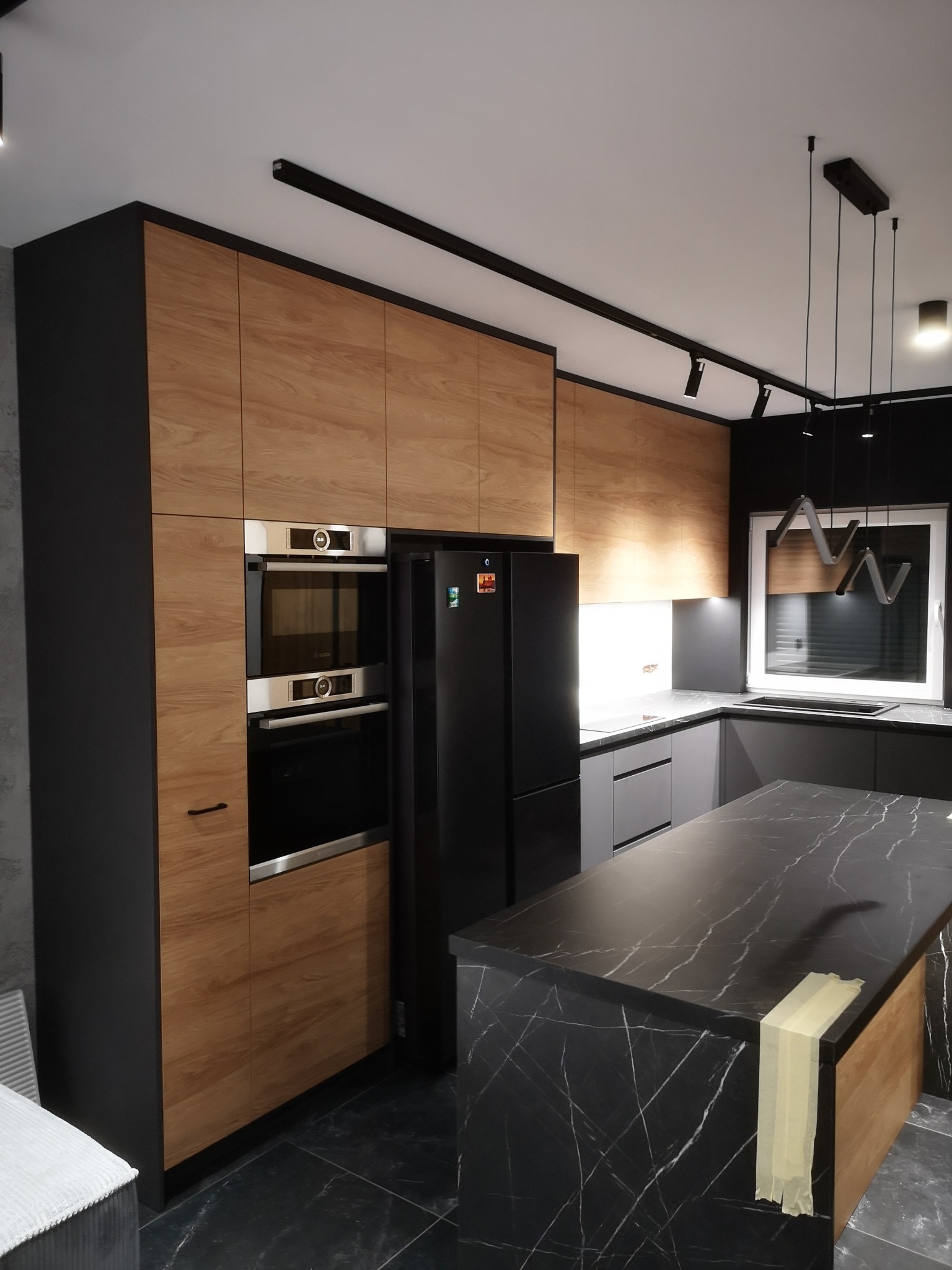 Nowoczesna kuchnia z zabudową w odcieniach drewna i czerni, wyspa z marmurowym blatem zabezpieczona taśmą malarską, oświetlenie szynowe i lampy wiszące.