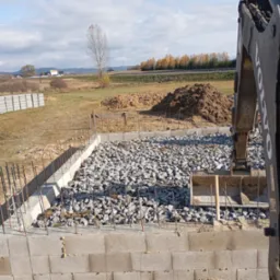 Budowa fundamentów: szalunek z bloczków betonowych, wypełniony kamieniem, wystające pręty zbrojeniowe, w tle koparka Volvo i hałdy ziemi.