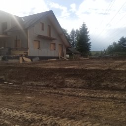 MAR-KOP Marcin Słodyczka - Dom w trakcie budowy, widoczne przygotowane podłoże z ubitej ziemi, elementy konstrukcyjne szkieletu, częściowo otynkowana elewacja, dach pokryty dachówką, w tle drzewa i pochmurne niebo.