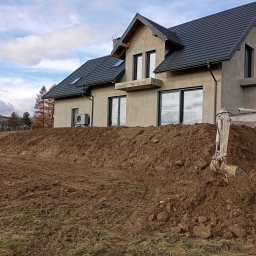 MAR-KOP Marcin Słodyczka - Nowo wybudowany dom z szarą elewacją i ciemną dachówką, widoczny z perspektywy wykopu, obok stoi mała koparka, w tle jesienny krajobraz wzgórz.