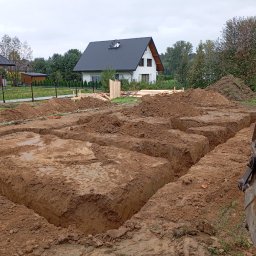 MAR-KOP Marcin Słodyczka - Wykopane rowy pod fundamenty domu jednorodzinnego, widoczna łyżka koparki, w tle budynki mieszkalne i drzewa.