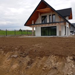 MAR-KOP Marcin Słodyczka - Nowoczesny dom z drewnianymi elementami w trakcie budowy, widoczny wykop pod przyszły ogród i ogrodzenie w tle.