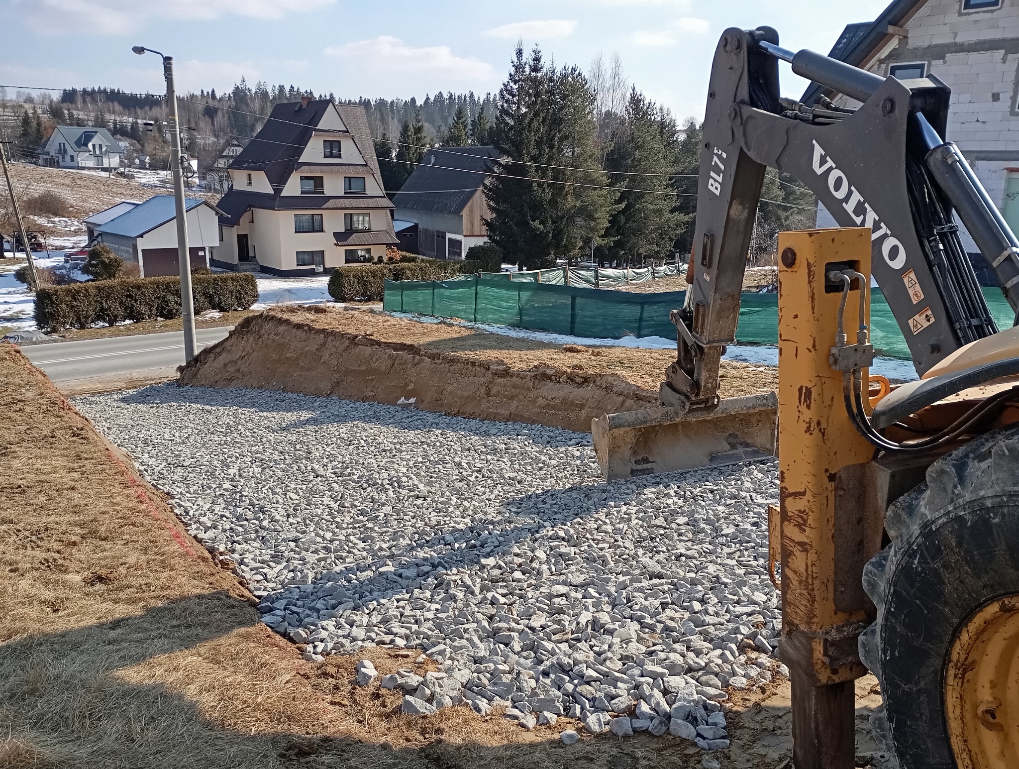 Żwirowe utwardzenie terenu pod budowę, widoczna koparka Volvo BL71, w tle domy jednorodzinne i drzewa.