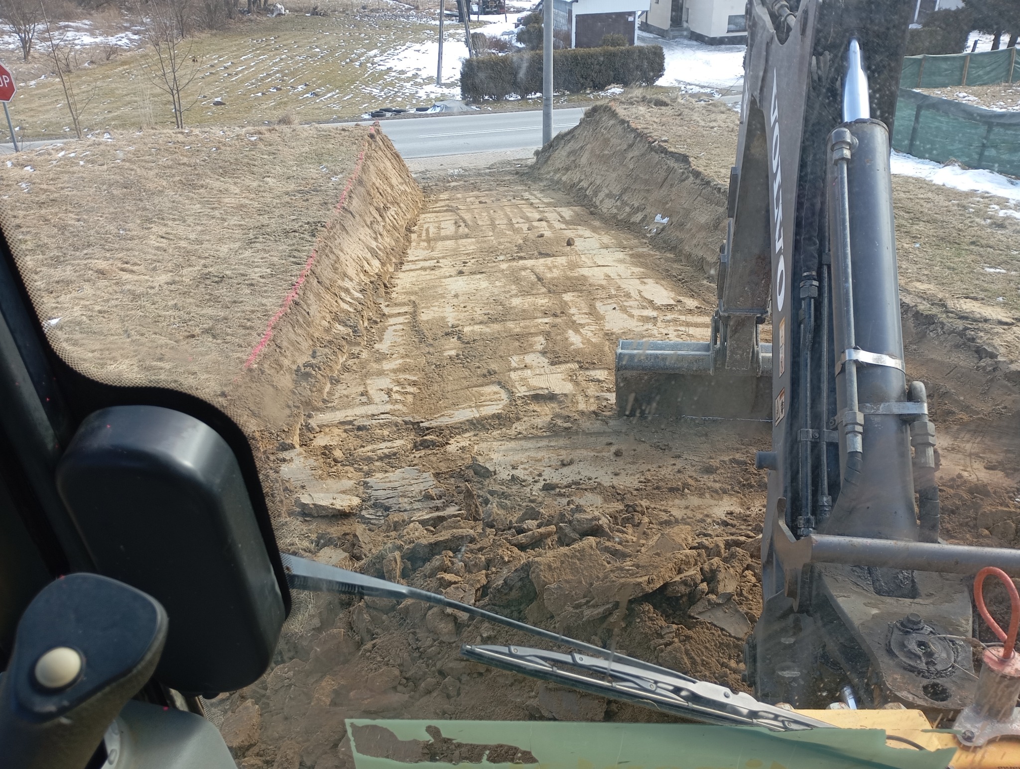 Widok z kabiny koparki na wykopany wąski rów w ziemi, widoczne ślady opon i fragmenty gruzu, tło stanowi droga i zabudowania.