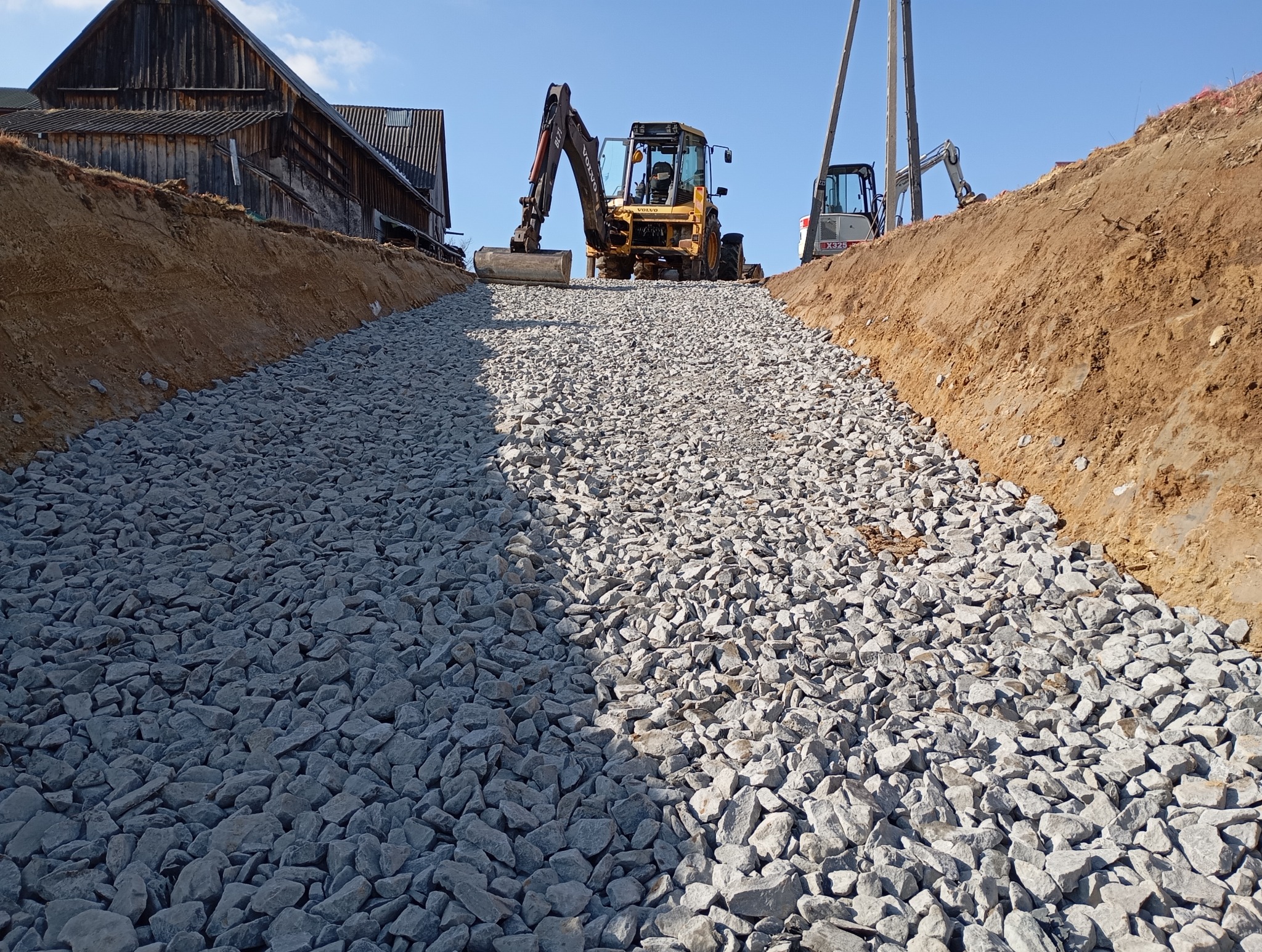 Żwirowa droga w wykopie, z widocznym żółtym ciągnikiem Volvo BL71 i koparką X325 w tle, stara drewniana stodoła po lewej stronie.