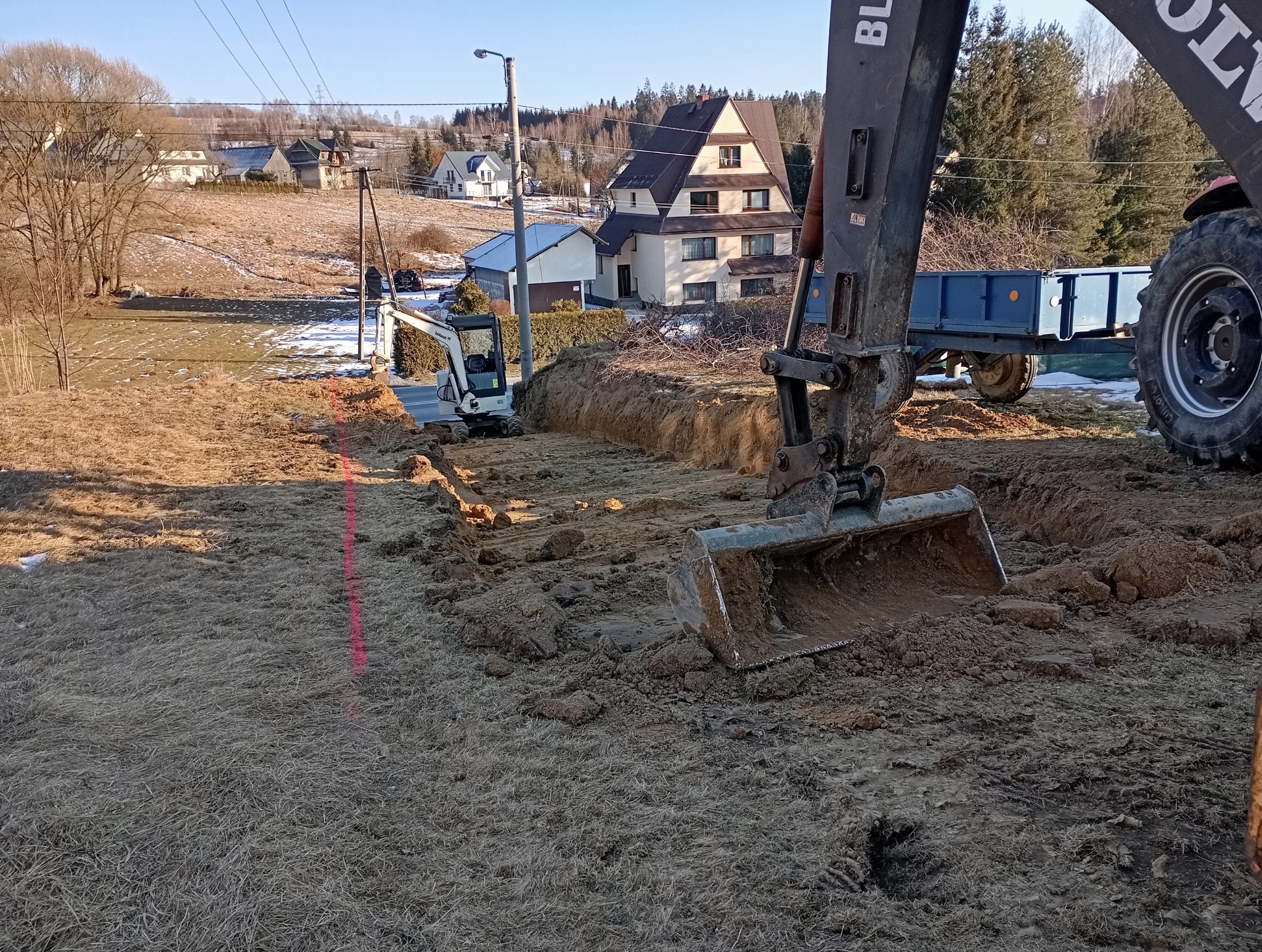 Dwa traktory podczas prac ziemnych na działce z widocznymi budynkami mieszkalnymi w tle, wyznaczona linia wykopu.