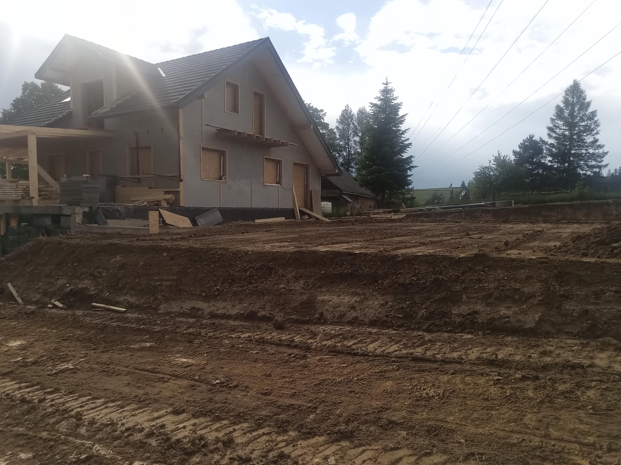 Dom w trakcie budowy, widoczne przygotowane podłoże z ubitej ziemi, elementy konstrukcyjne szkieletu, częściowo otynkowana elewacja, dach pokryty dachówką, w tle drzewa i pochmurne niebo.