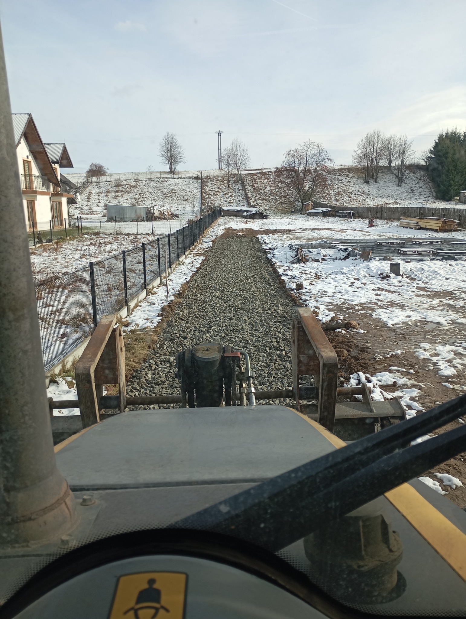 Widok z kabiny maszyny budowlanej na utwardzoną, żwirową drogę w zimowym krajobrazie, z widoczną zabudową jednorodzinną i ogrodzeniem w tle.