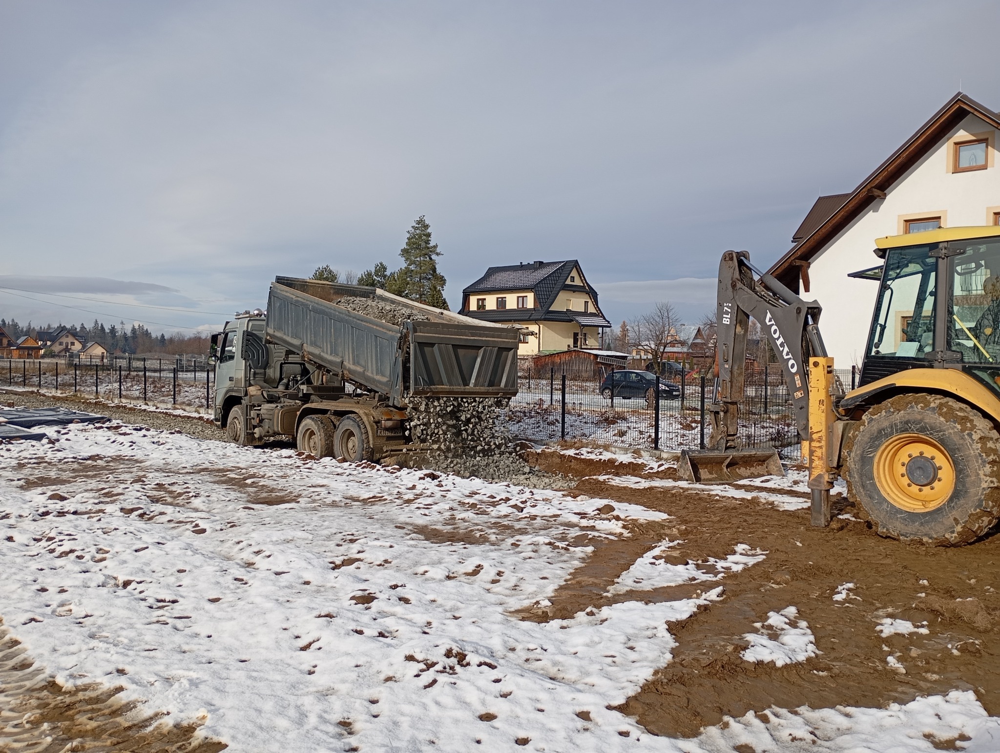 Ciężarówka wyładowuje gruz na placu budowy, obok stoi żółta koparko-ładowarka Volvo BL71, w tle widoczne domy i częściowo ośnieżony teren.