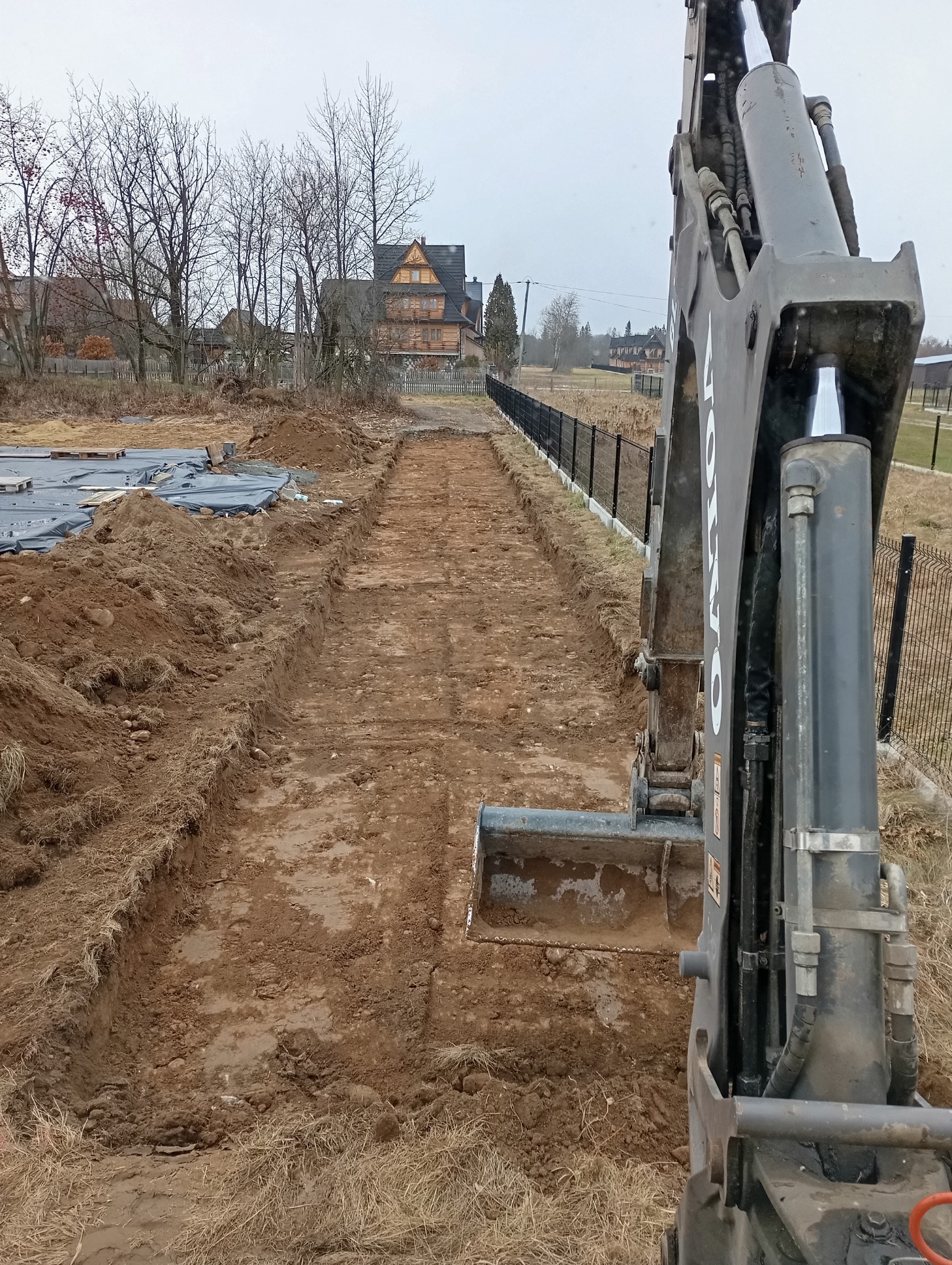 Koparka Volvo podczas wykonywania wykopu w ziemi, widoczny krajobraz z domami w stylu góralskim w tle.