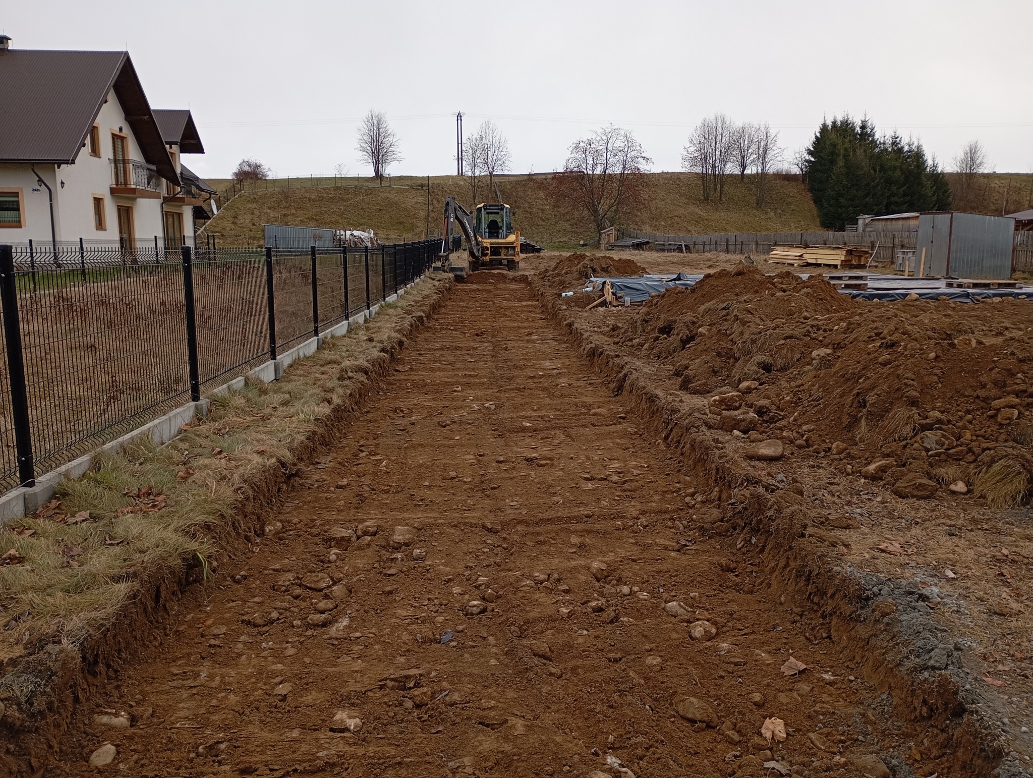 Żółta koparka Volvo na placu budowy, przygotowująca teren pod przyszłą drogę, widoczne świeżo wykopane rowy i pryzmy ziemi, w tle dom jednorodzinny i drzewa.