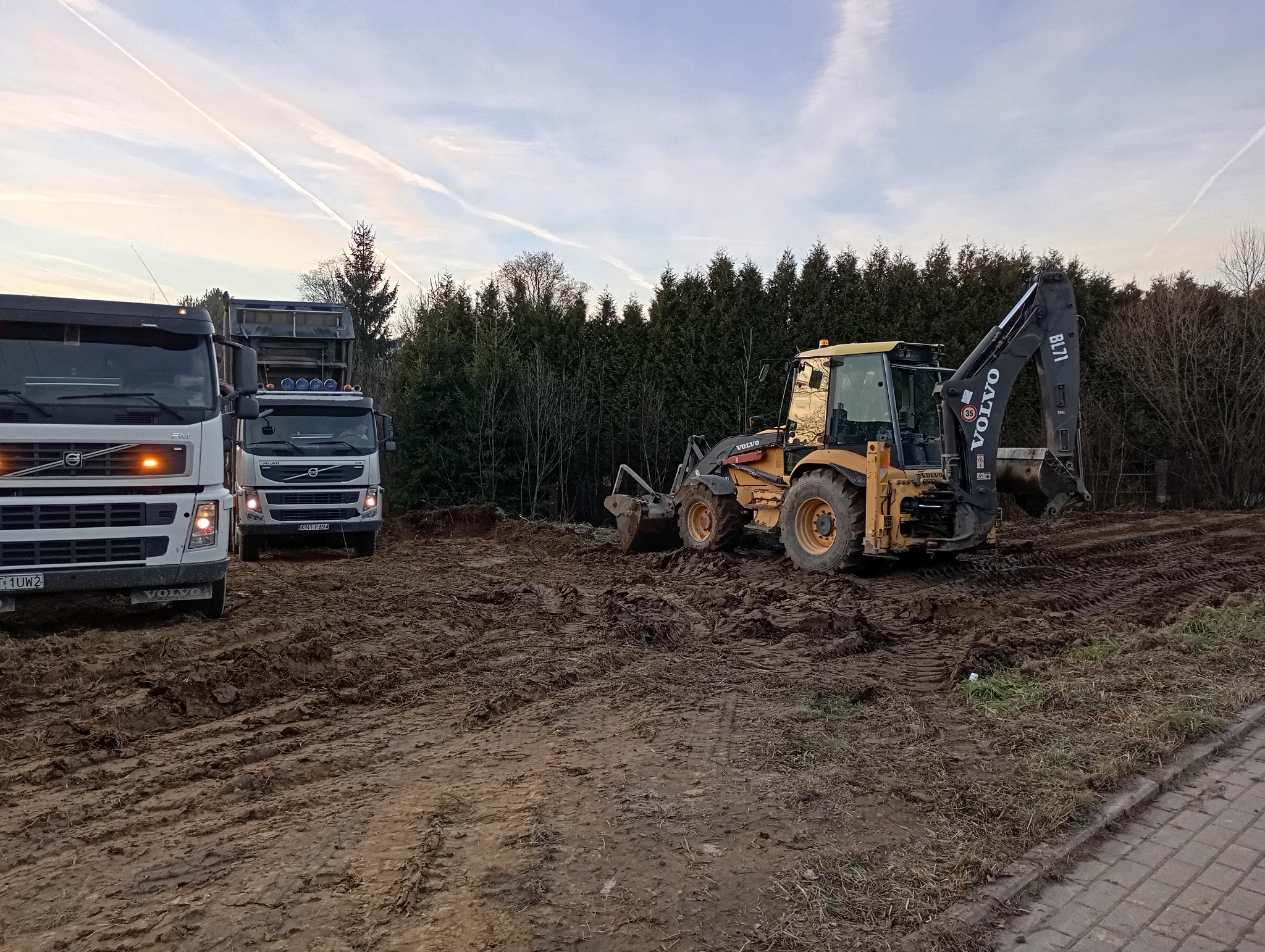 Dwa samochody ciężarowe Volvo i koparko-ładowarka Volvo BL71 na placu budowy z ubitym błotem, w tle drzewa iglaste i smugi na niebie.