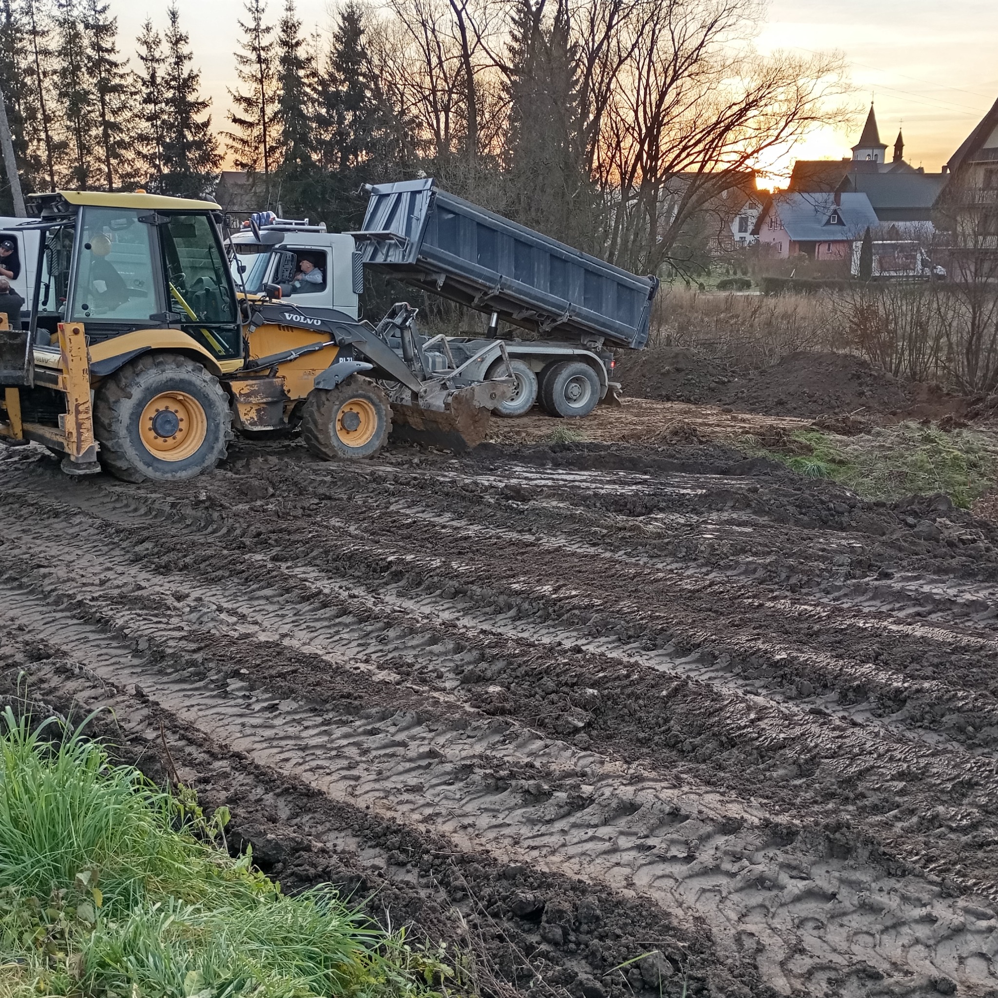Żółta ładowarka Volvo z łyżką i wywrotka w trakcie prac ziemnych na placu budowy, widoczne ślady opon w błocie, w tle budynki i zachodzące słońce.