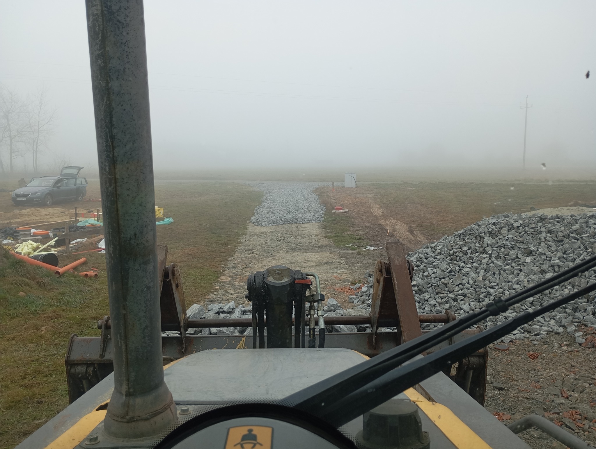 Widok z kabiny ładowarki na plac budowy w mglisty dzień, widoczny nasyp z kruszywa, rury, samochód osobowy i elementy infrastruktury w oddali.