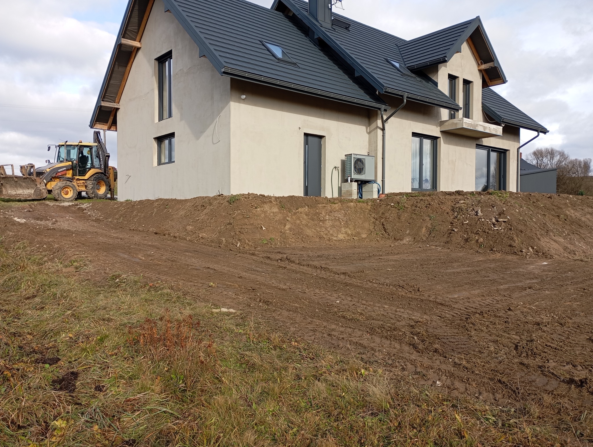 Nowy dom w trakcie budowy z widocznym traktorem Volvo na tle, przygotowującym teren pod ogród.
