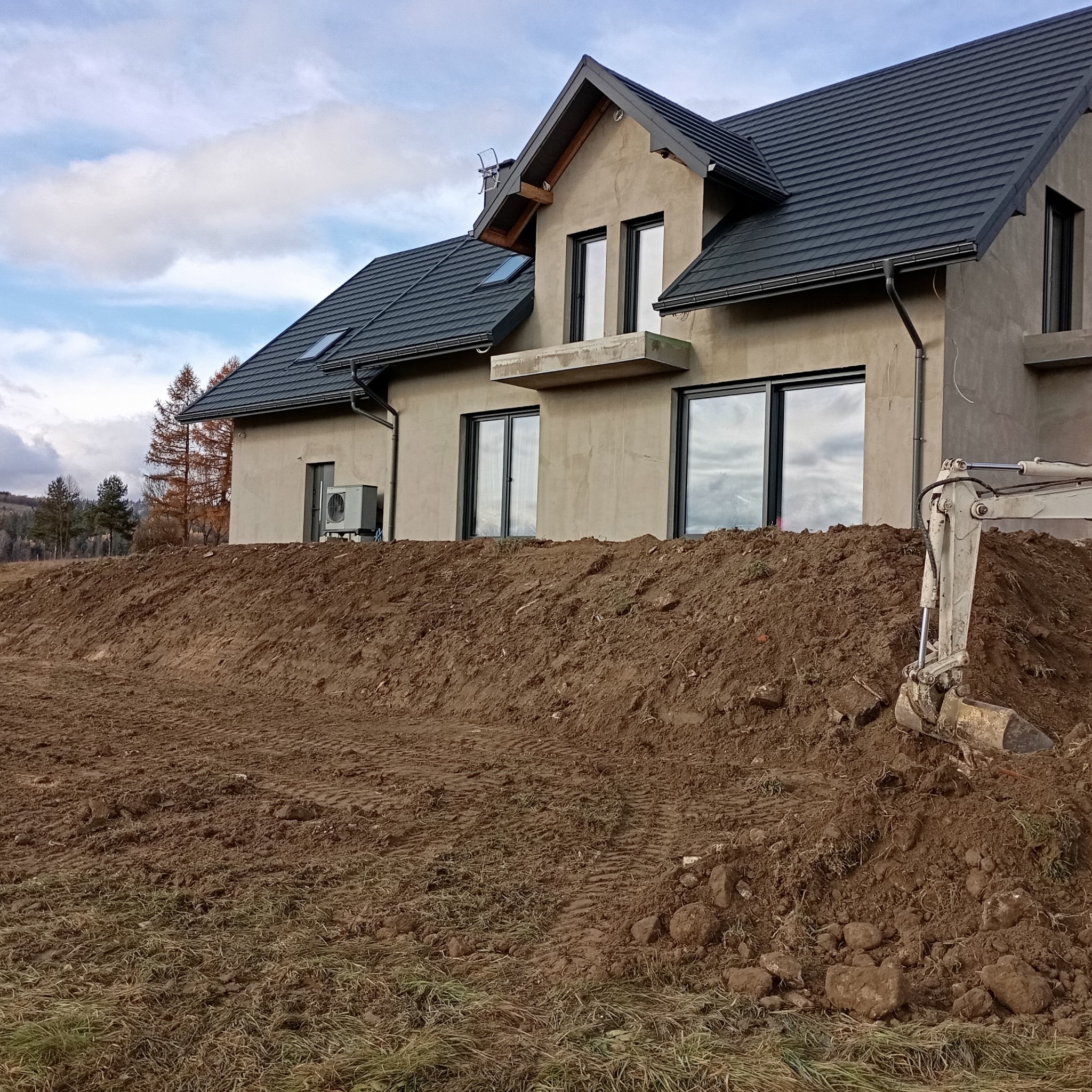 Nowo wybudowany dom z szarą elewacją i ciemną dachówką, widoczny z perspektywy wykopu, obok stoi mała koparka, w tle jesienny krajobraz wzgórz.