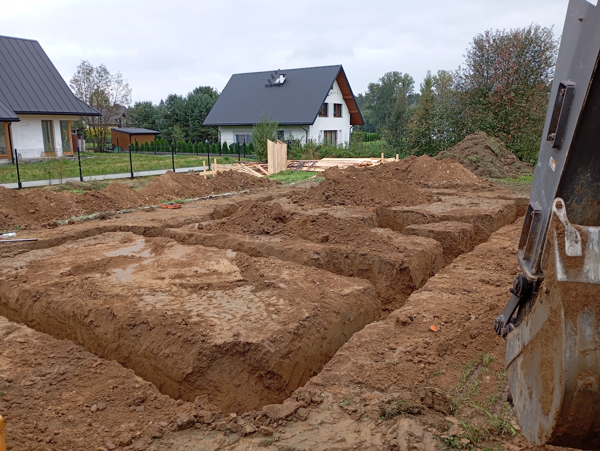 Wykopane rowy pod fundamenty domu jednorodzinnego, widoczna łyżka koparki, w tle budynki mieszkalne i drzewa.