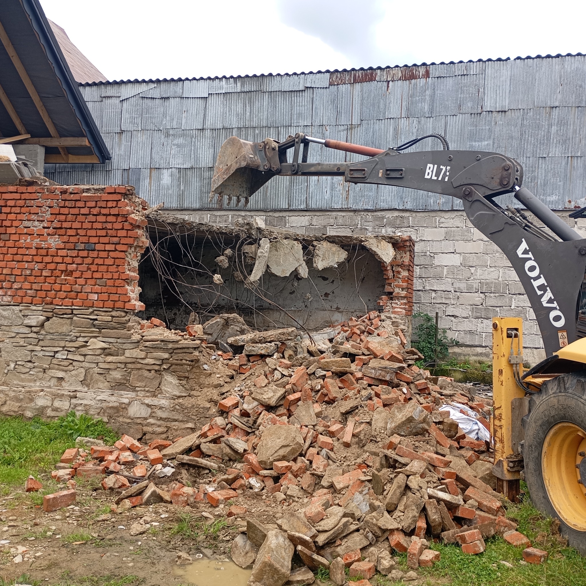 Żółta ładowarka Volvo BL71 z łyżką na wysięgniku, w trakcie rozbiórki muru z czerwonej cegły i kamienia, z gruzem na ziemi i blaszakiem w tle.