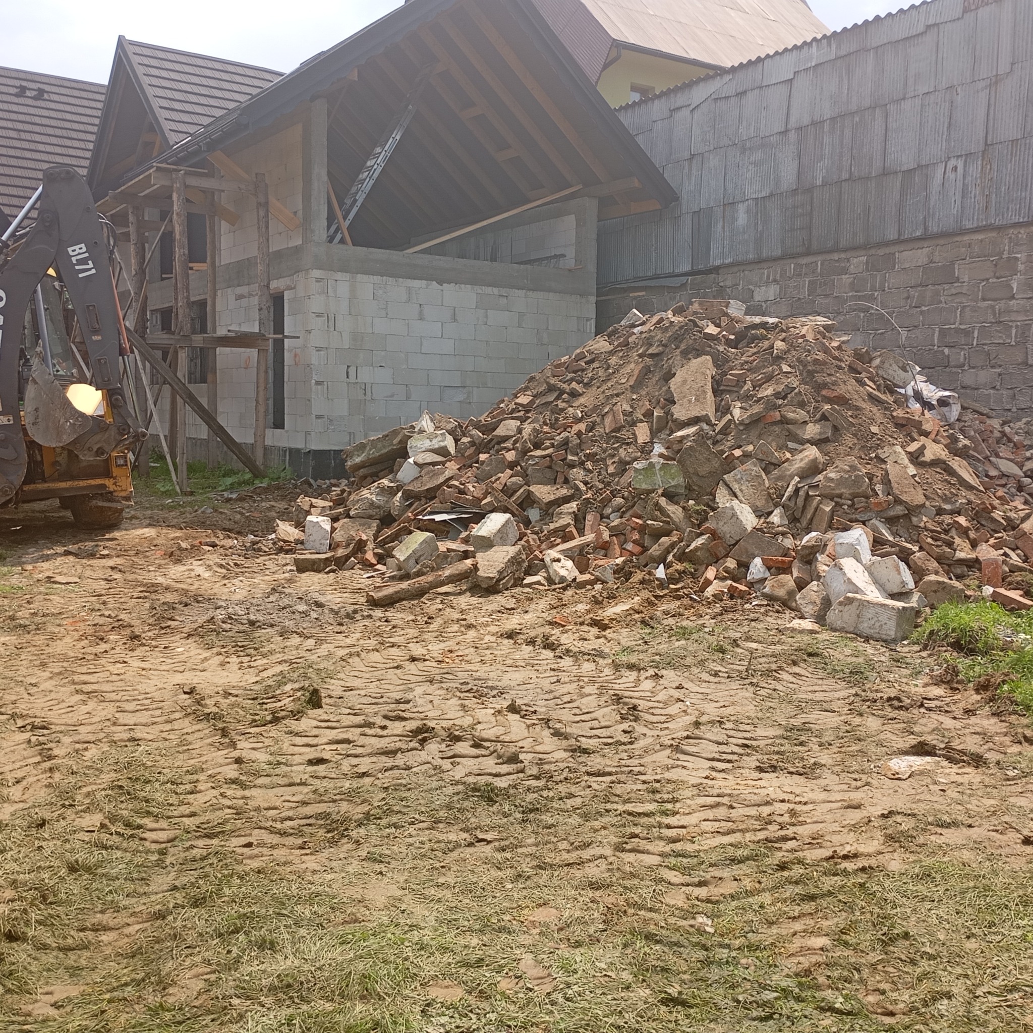 Żółta koparka obok budowanego domu z białego pustaka, przed nią pryzma gruzu i cegieł na ubitej ziemi z wyraźnymi śladami opon.
