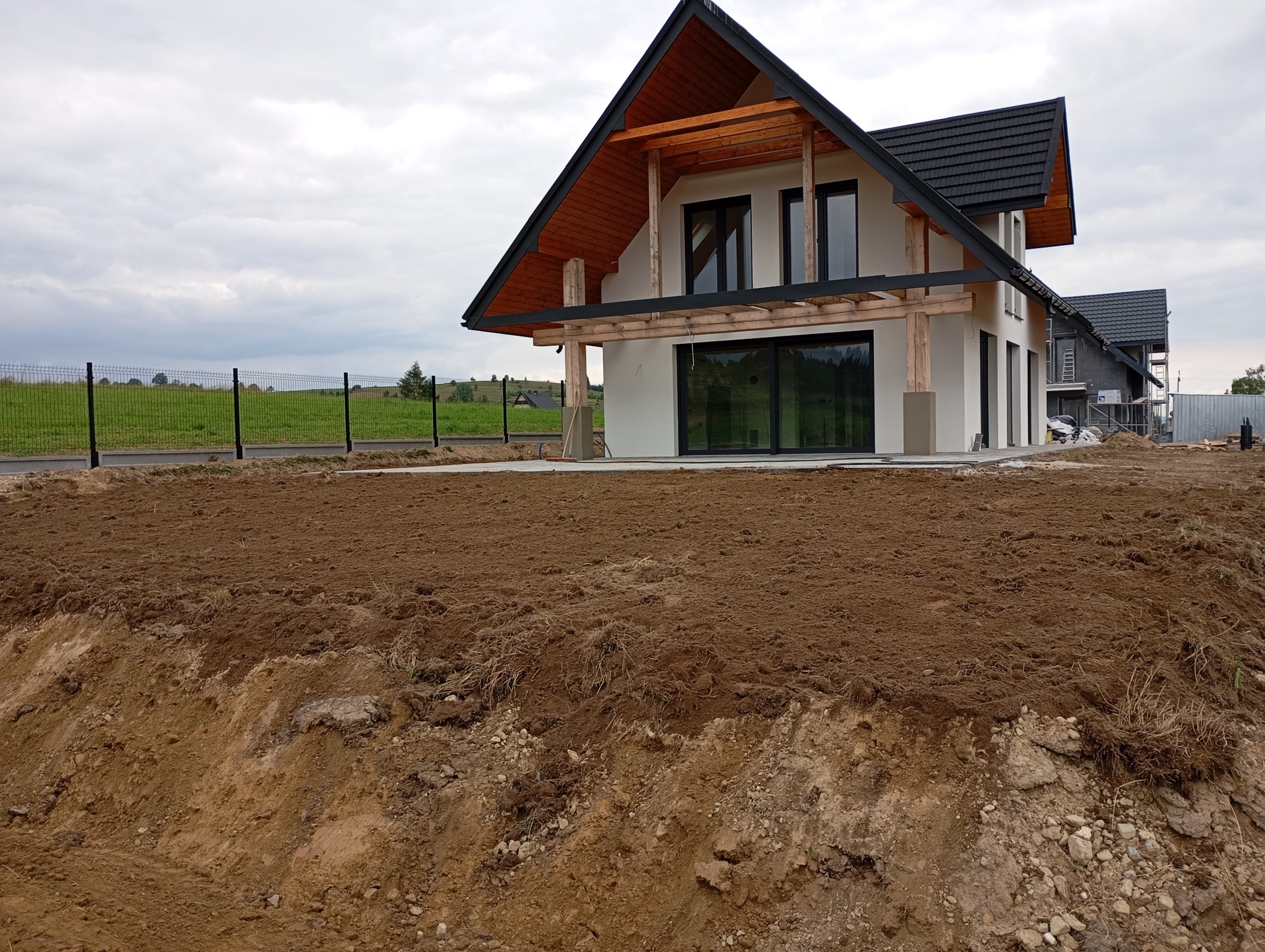 Nowoczesny dom z drewnianymi elementami w trakcie budowy, widoczny wykop pod przyszły ogród i ogrodzenie w tle.