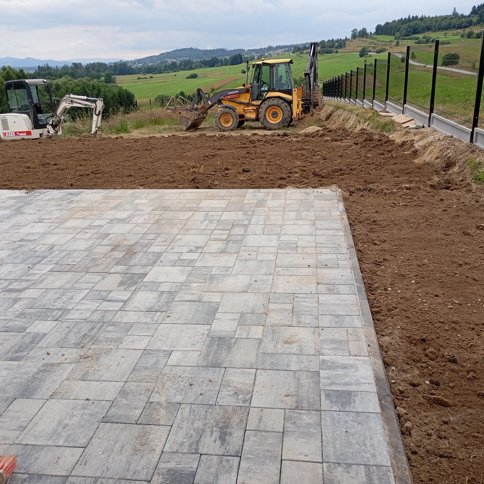 Ułożona szara kostka brukowa na przygotowanym terenie z widocznymi maszynami budowlanymi w tle i nowym ogrodzeniem.
