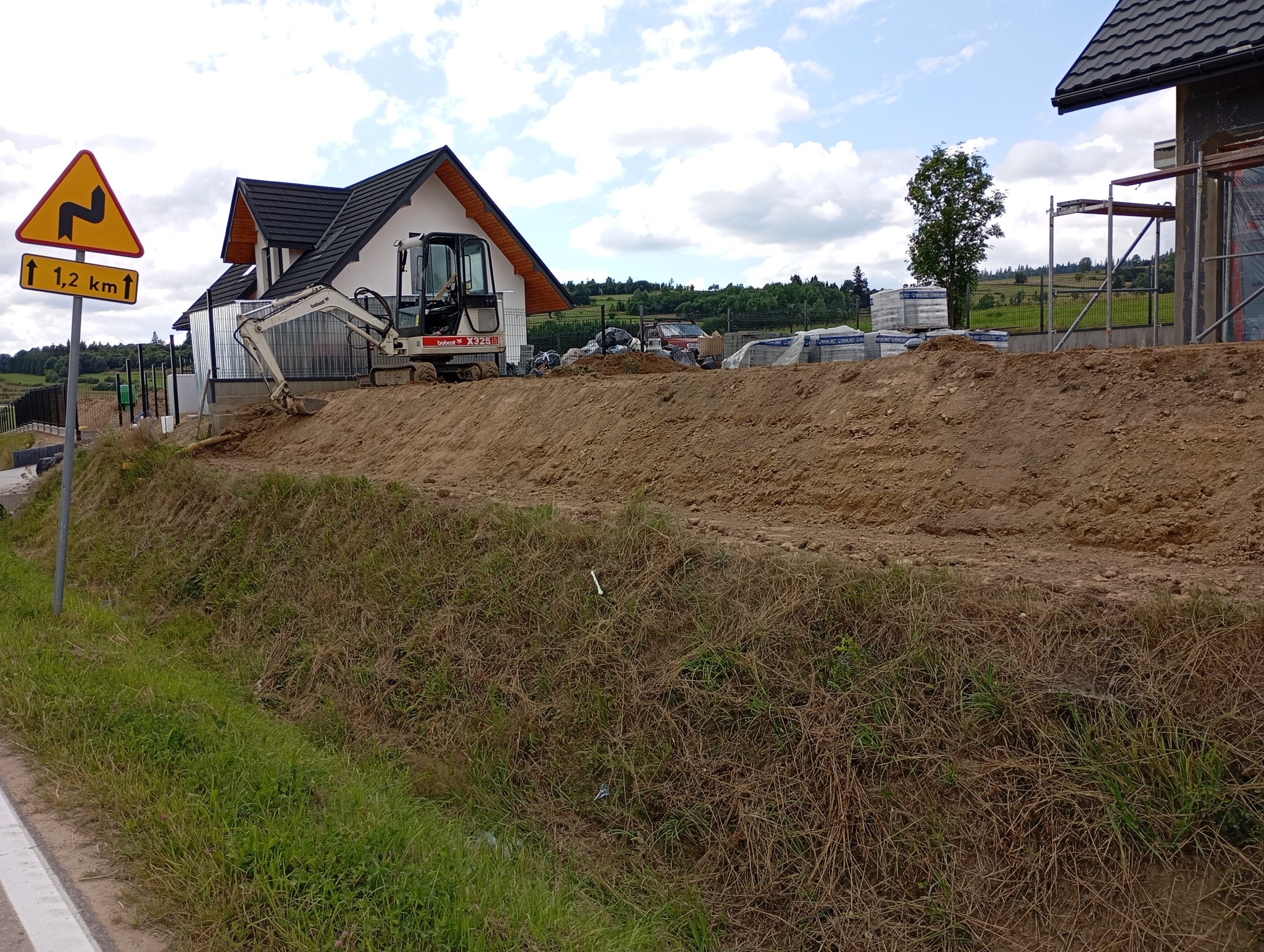 Biała mini koparka Bobcat X325 na wzgórzu z ziemi, w tle budowa domu z czarnym dachem i stosem bloczków, po lewej znak drogowy ostrzegający o zakręcie.