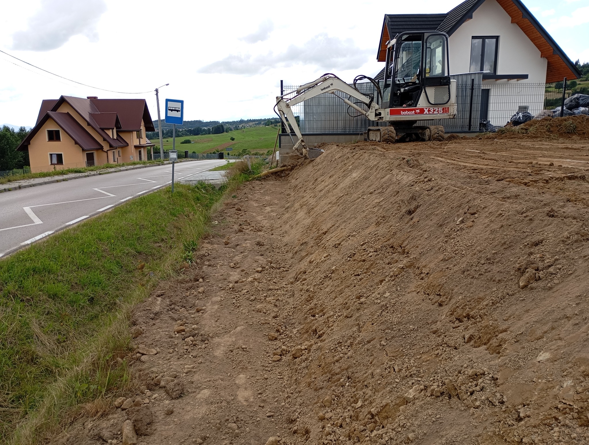 Mini koparka Bobcat X325 na gruncie obok nowo wybudowanego domu z szarą elewacją i czarnym dachem, widoczny wykop ziemi i droga z przystankiem autobusowym w tle.