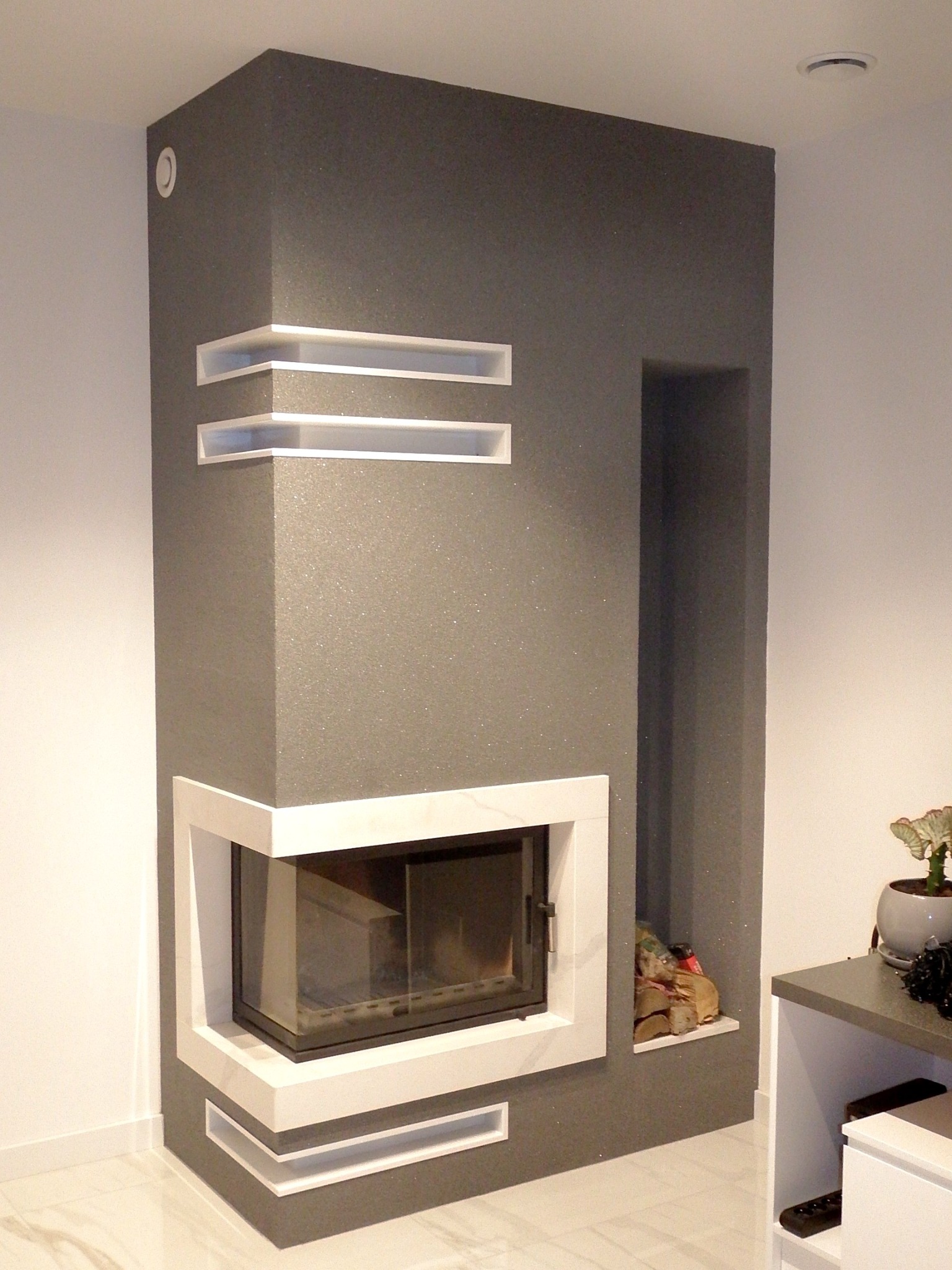 Nowoczesny kominek narożny z szarą, błyszczącą obudową, białymi ramami i wnękami, z drewnem ułożonym w bocznej wnęce, na tle białej ściany i marmurowej podłogi.