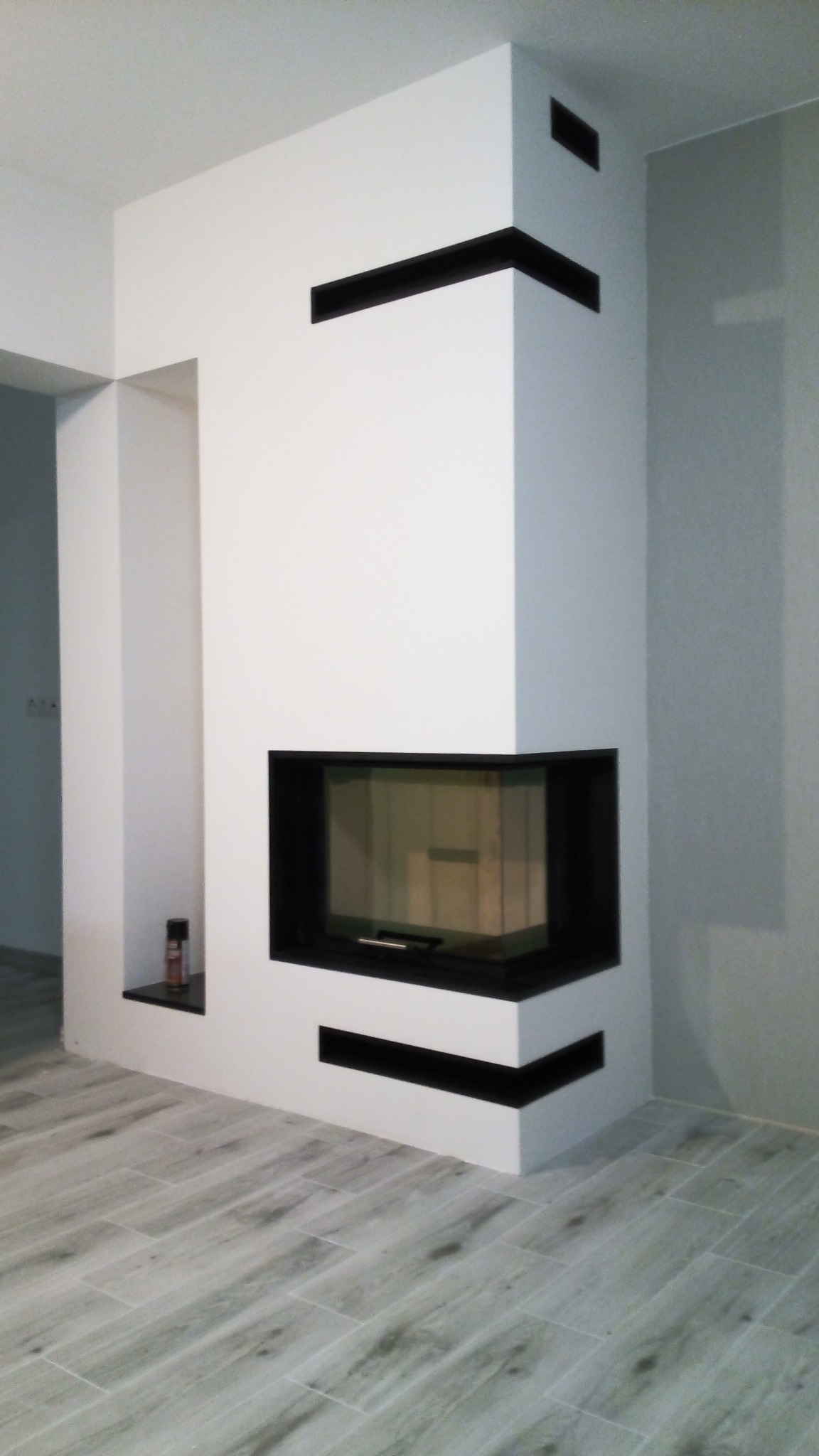 Nowoczesny, narożny kominek w obudowie z białego gładkiego tynku z czarnymi akcentami, wnęka obok z czarną półką, na której stoi mała butelka, podłoga wyłożona płytkami imitującymi drewno.