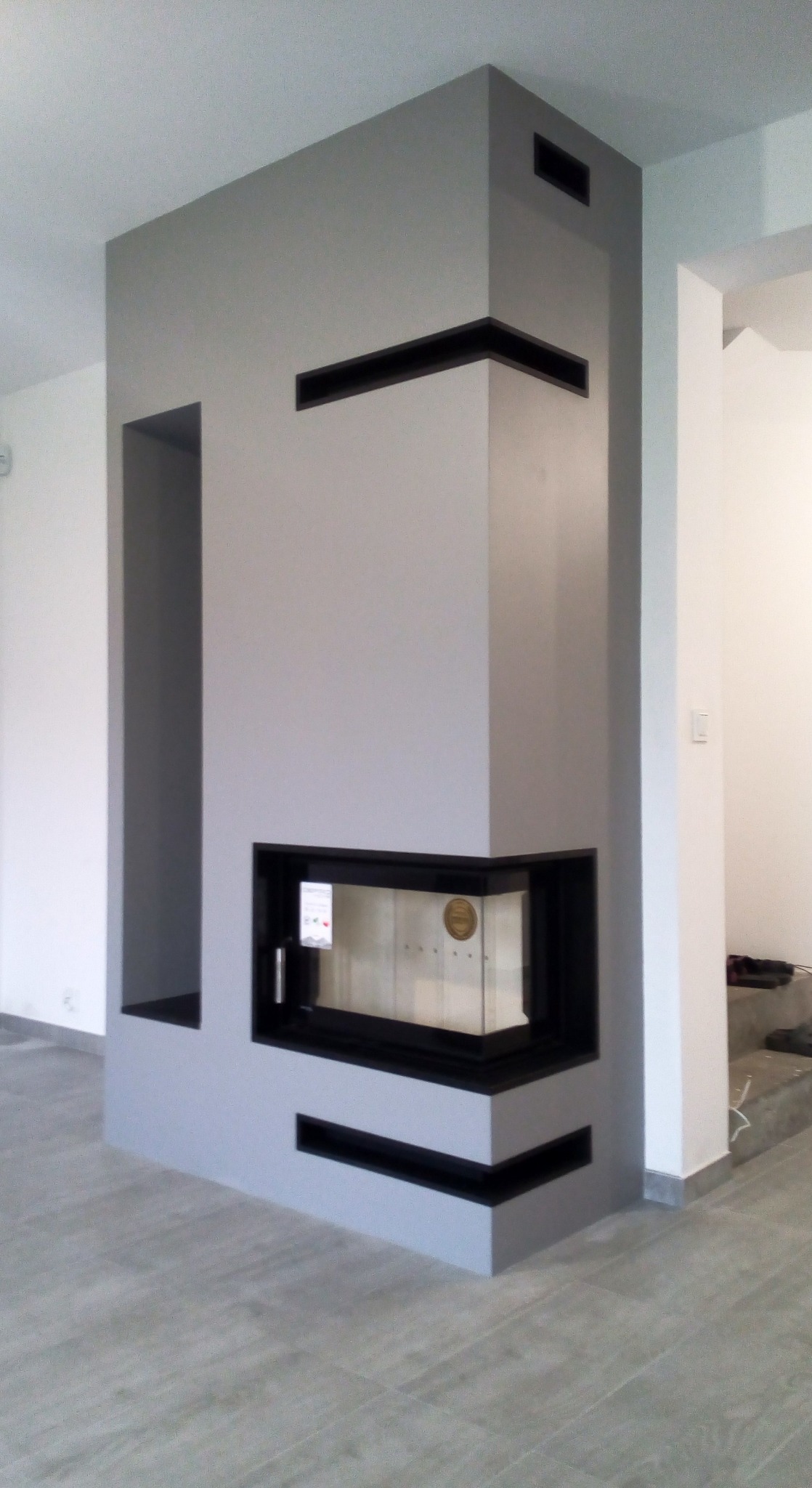 Nowoczesny, narożny wkład kominkowy z czarną ramą, obudowany szarą konstrukcją z wnękami, w minimalistycznym wnętrzu.