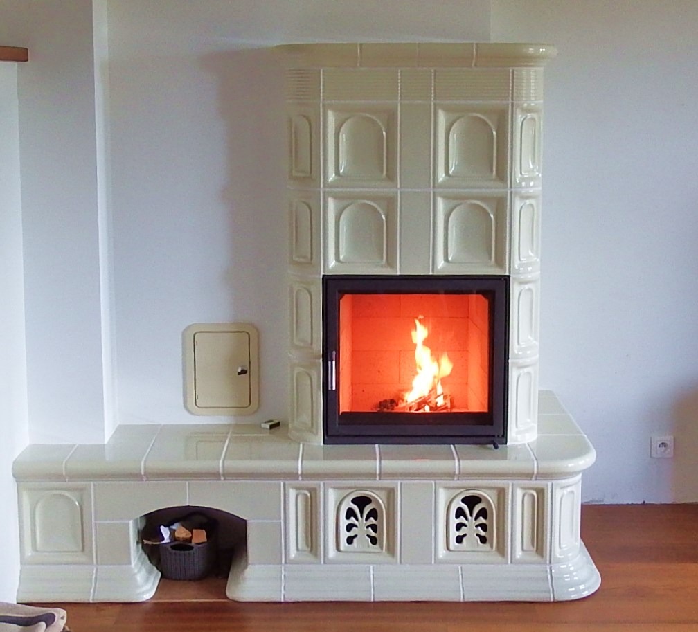 Biały, kaflowy piec z paleniskiem, płomienie widoczne przez szybę, drewno ułożone w dolnej wnęce, minimalistyczne wnętrze z drewnianą podłogą.